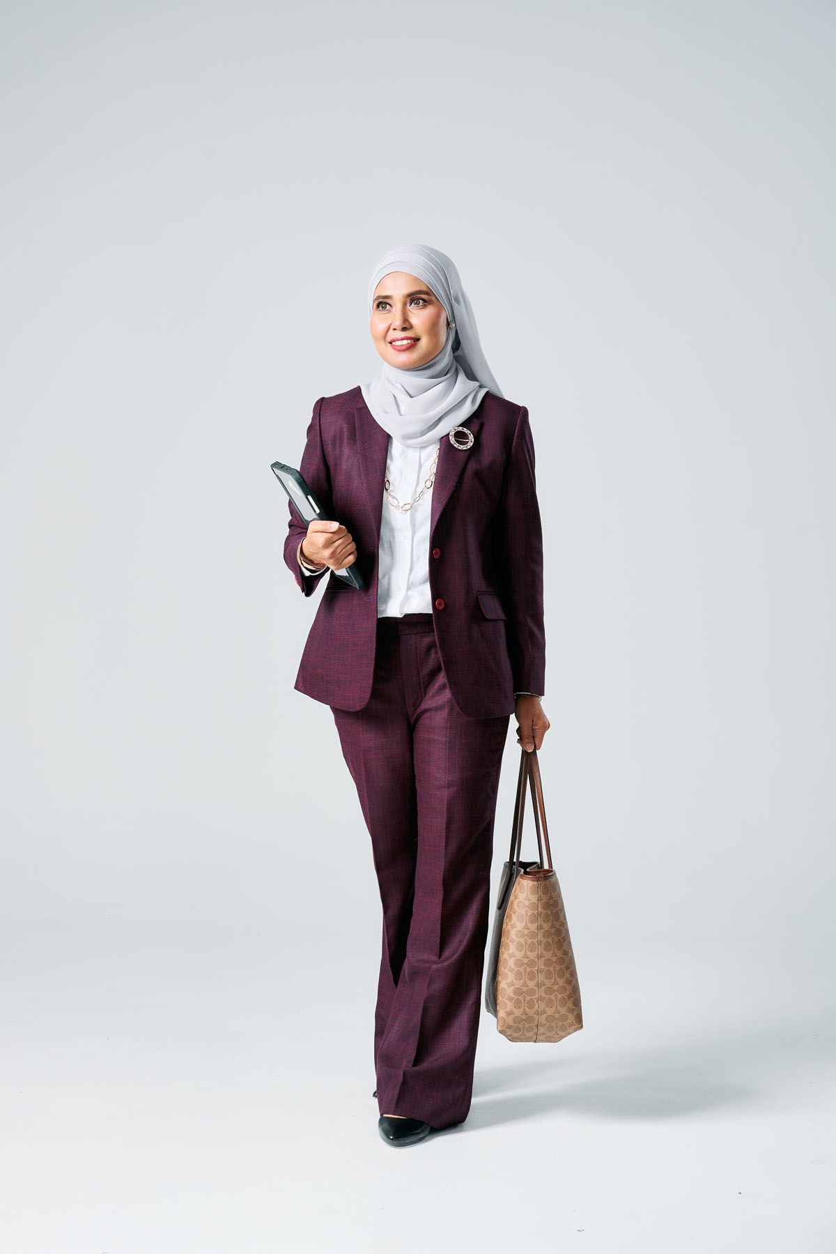 Professional Muslim woman in maroon suit and gray hijab walking while holding tablet and handbag.