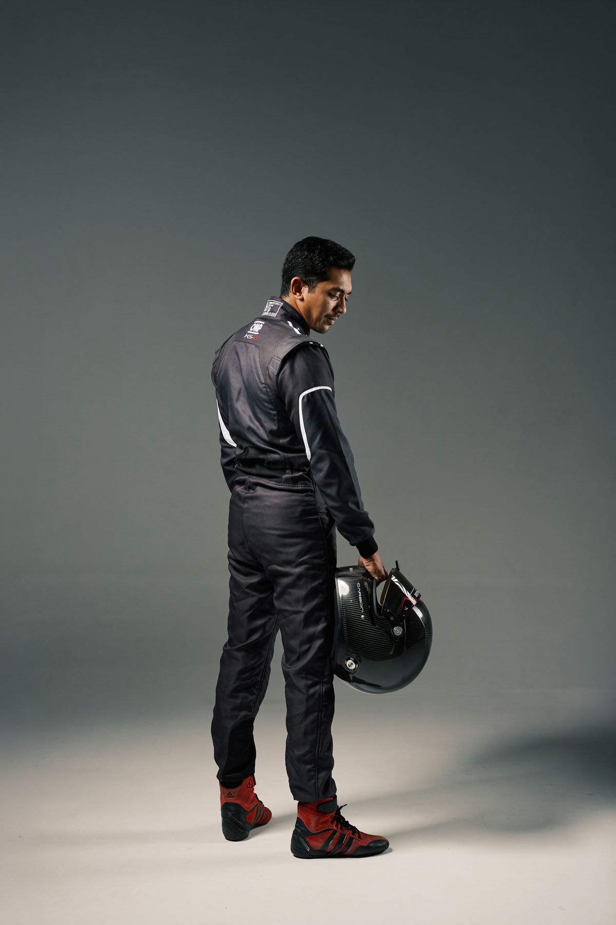 Race car driver in black racing suit holding helmet, standing in studio with gray background.