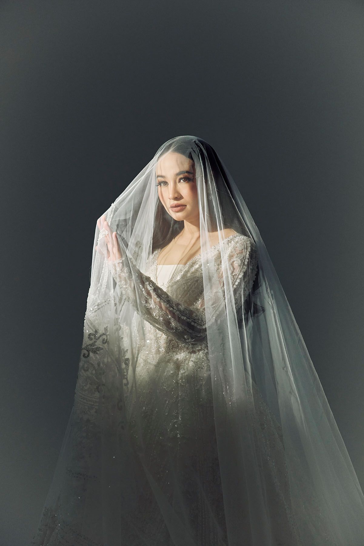 Elegant bride in a white lace wedding gown lifting a sheer veil over her face in a studio.