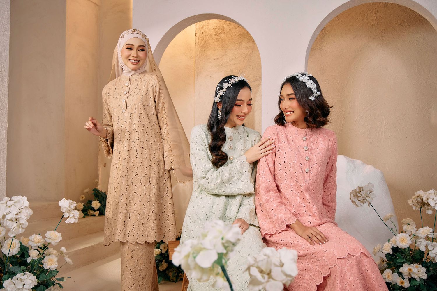 Three women smiling in pastel broderie anglaise baju kurung sets surrounded by white flowers indoors.