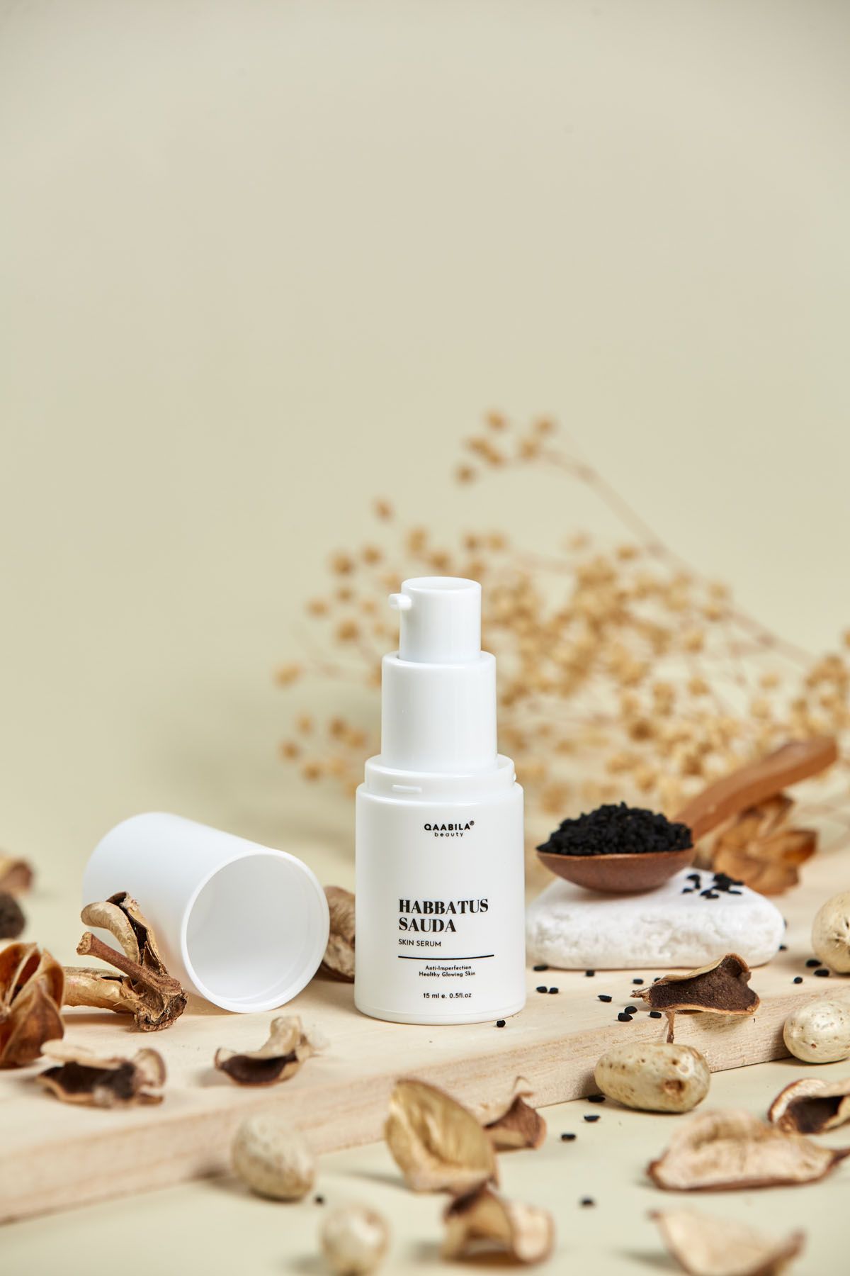 Qaabila Habbatus Sauda skin serum pump bottle with black seeds, dried botanicals on wooden board, beige background.