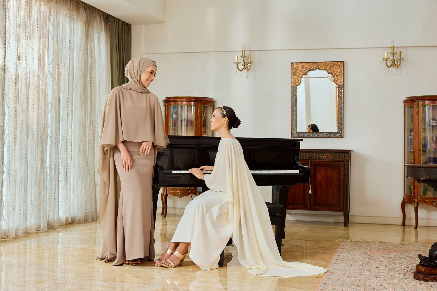Two women in modest elegant attire near a grand piano in a classic interior room.