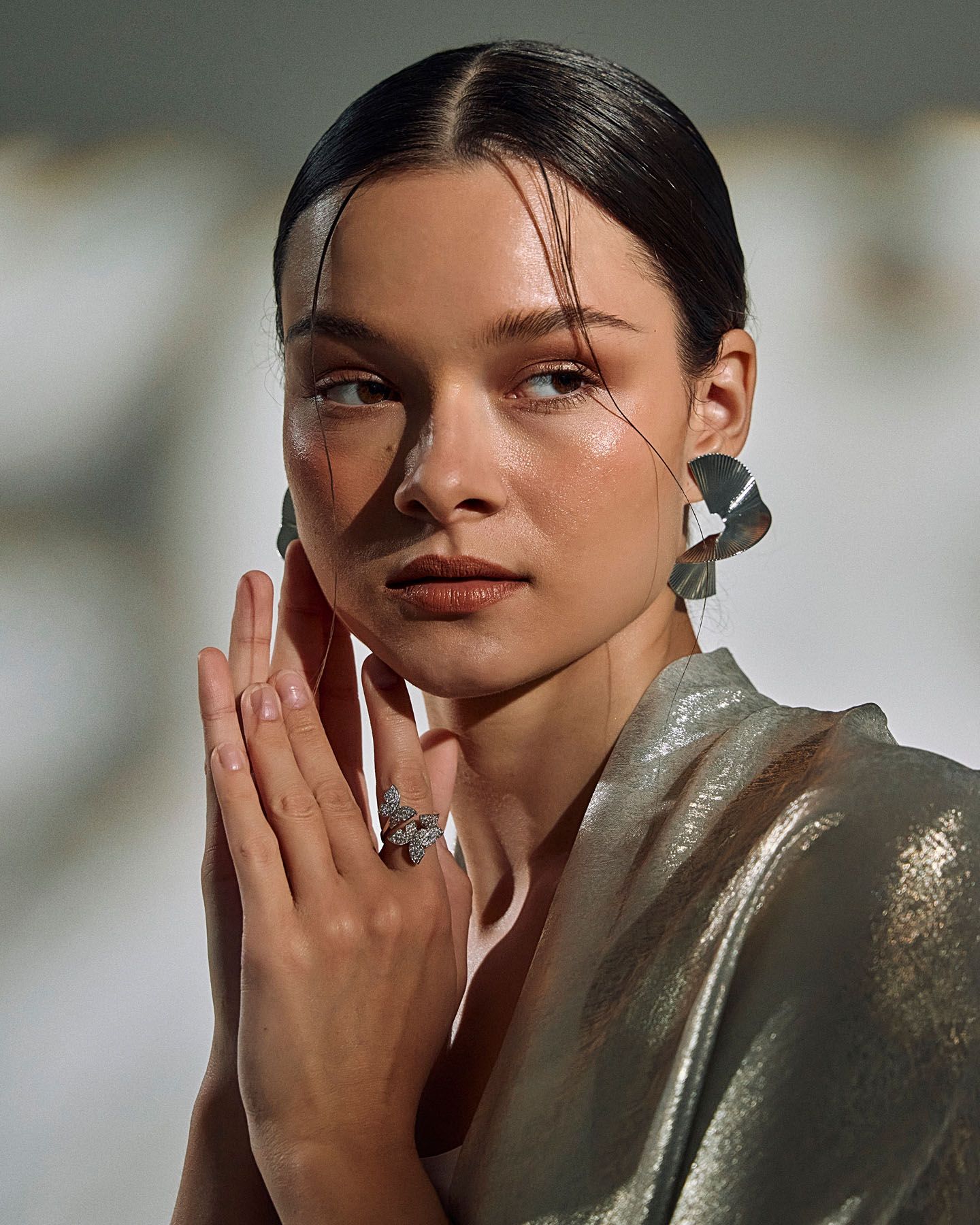Woman in silver metallic draped garment with statement earrings and ring prays hands together softly.
