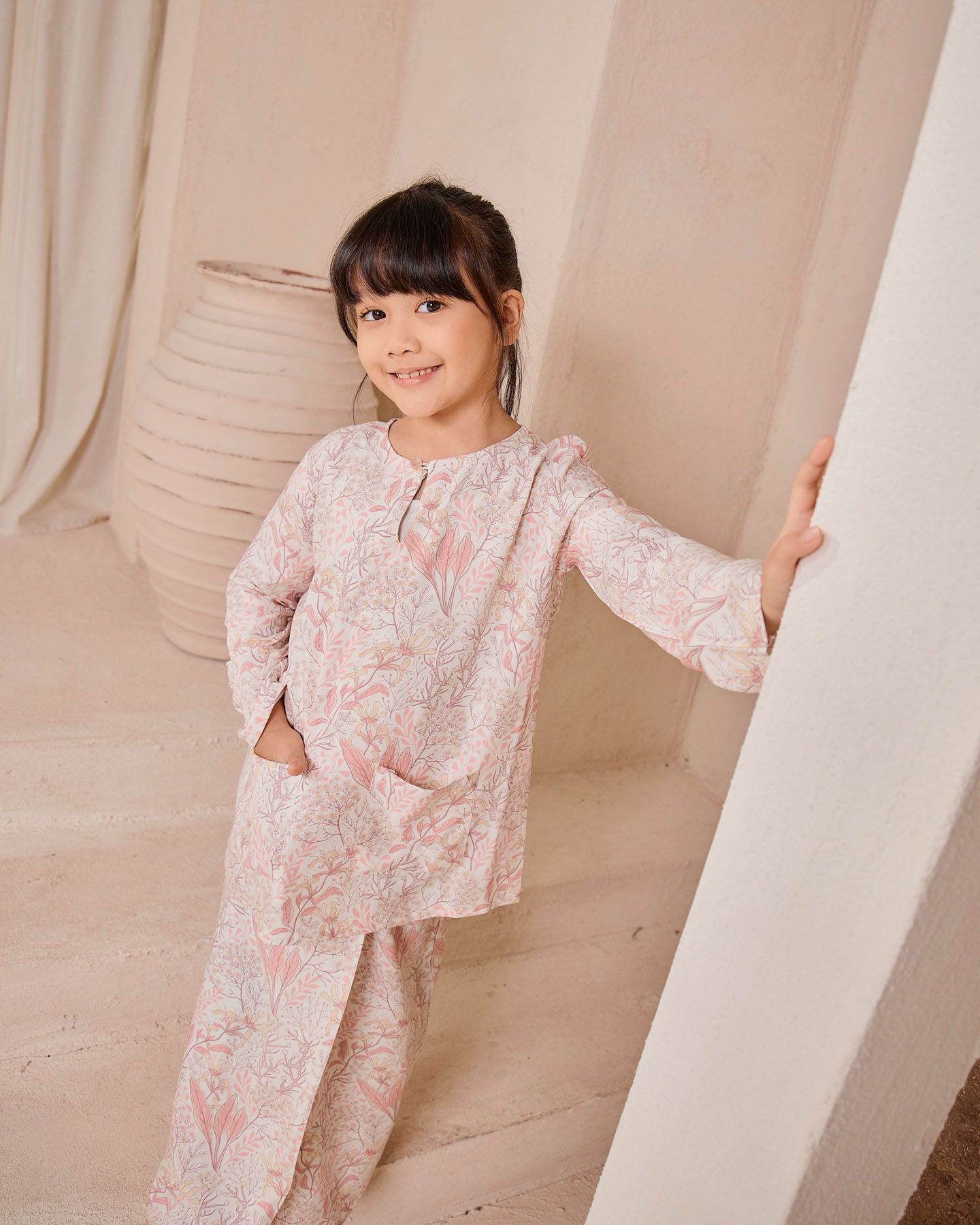 A smiling young girl in a pink floral baju kurung stands confidently in a warmly lit beige studio setting.