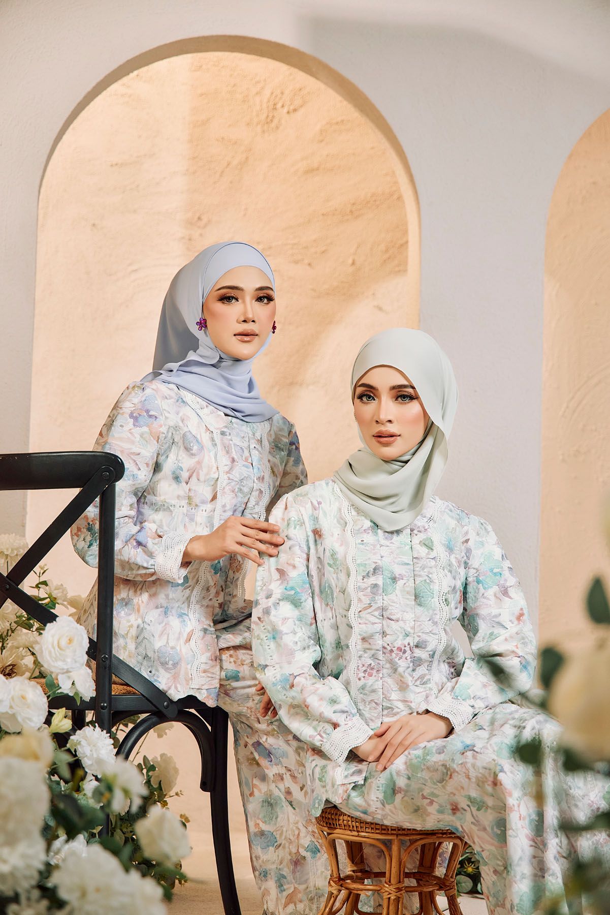 Two women in floral baju kurung with light blue hijabs pose near white flowers indoors.