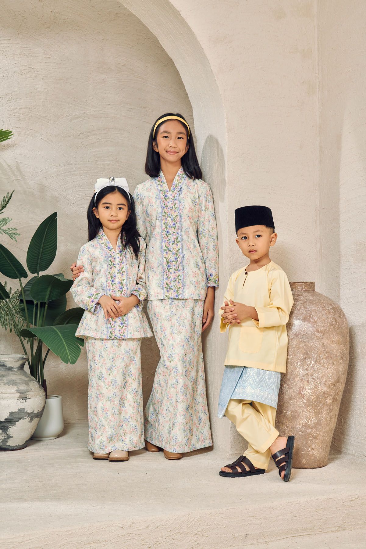 Three children in traditional Malay attire, two girls in floral baju kurung and boy in yellow baju melayu.