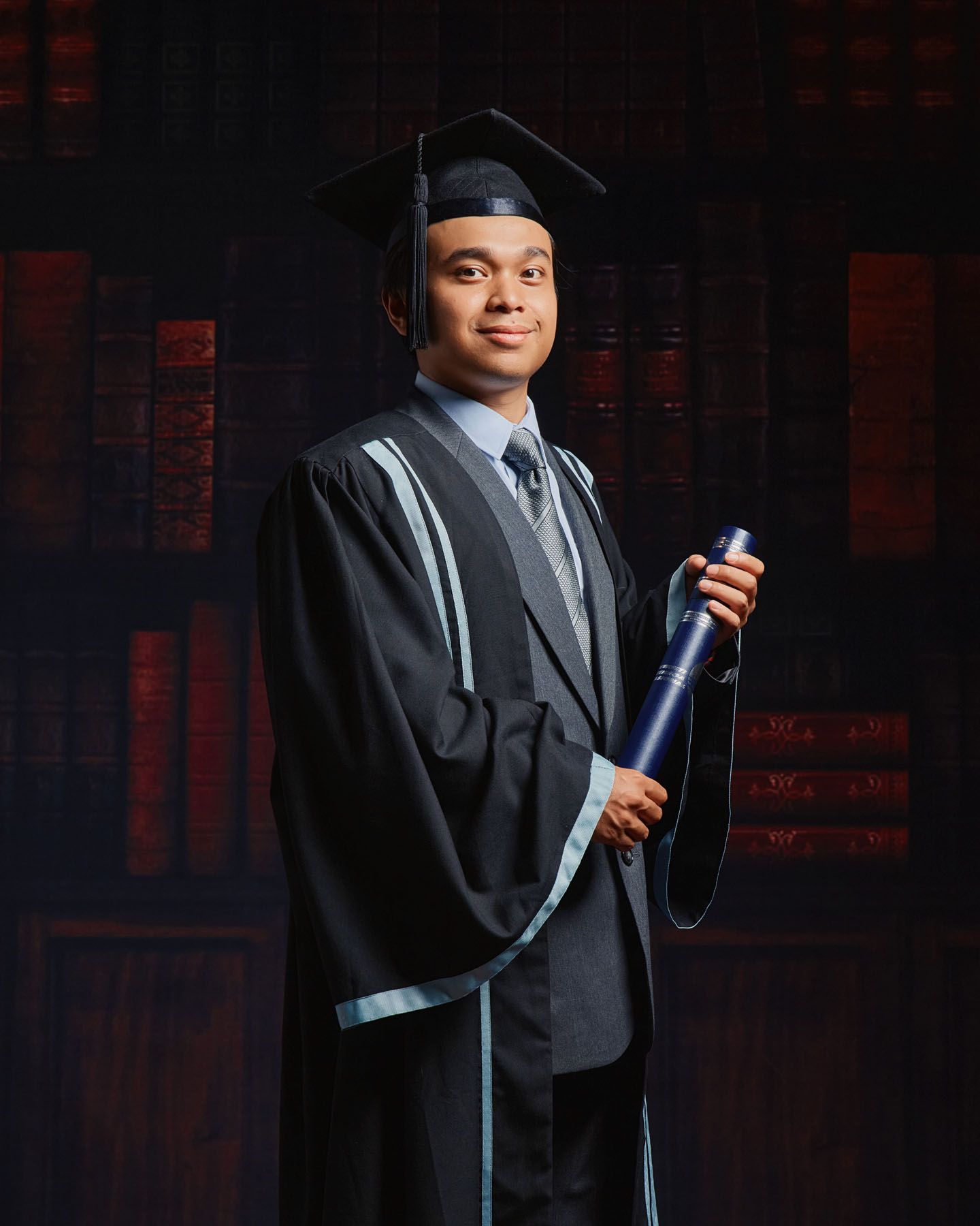 Young male graduate in black academic regalia holding diploma scroll posing in professional studio.