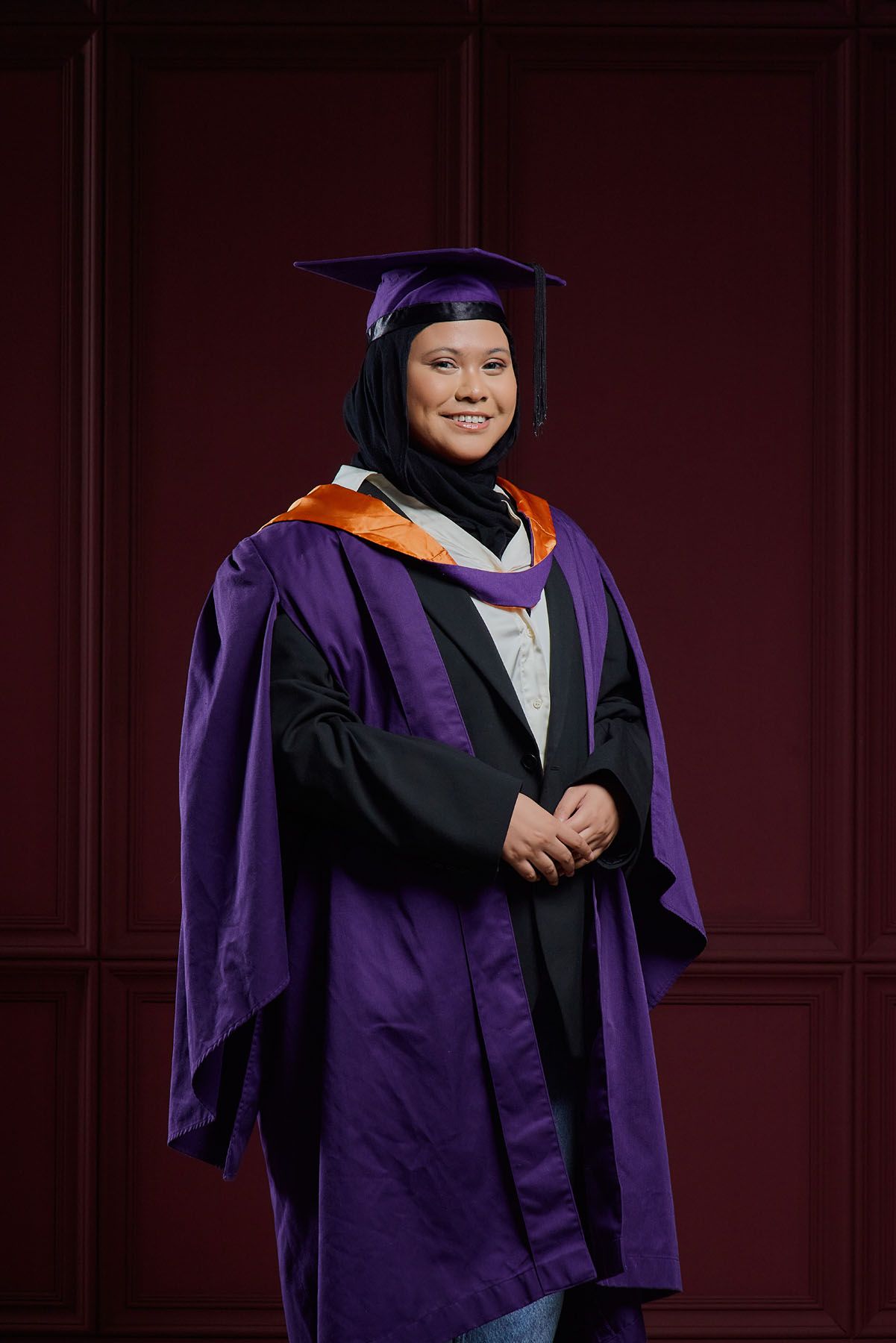 Muslim woman in purple graduation gown and mortarboard poses against dark red background.