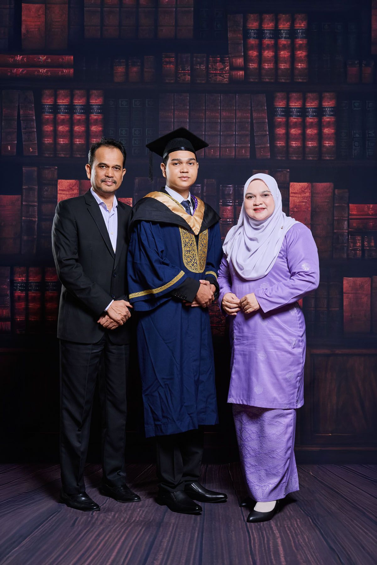 Graduate in blue convocation robe with parents, mother in purple baju kurung and hijab, against library backdrop.