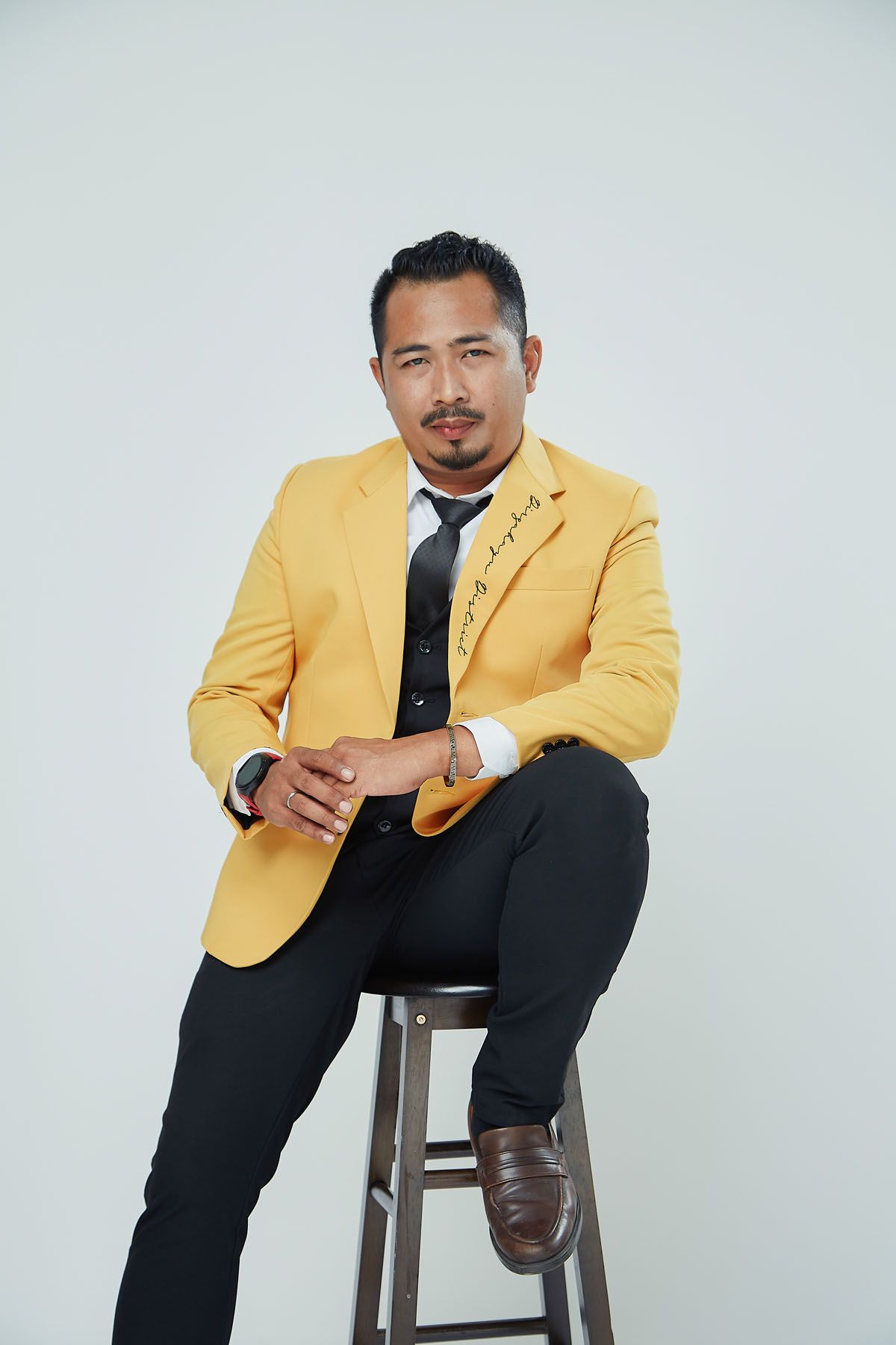 Professional Asian man in yellow blazer and black tie sitting on wooden stool with clasped hands.