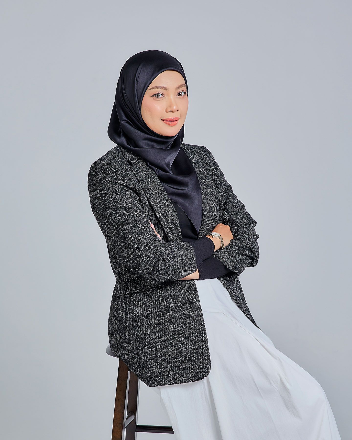 Muslim woman in gray herringbone blazer and navy hijab sitting on stool with arms crossed confidently.