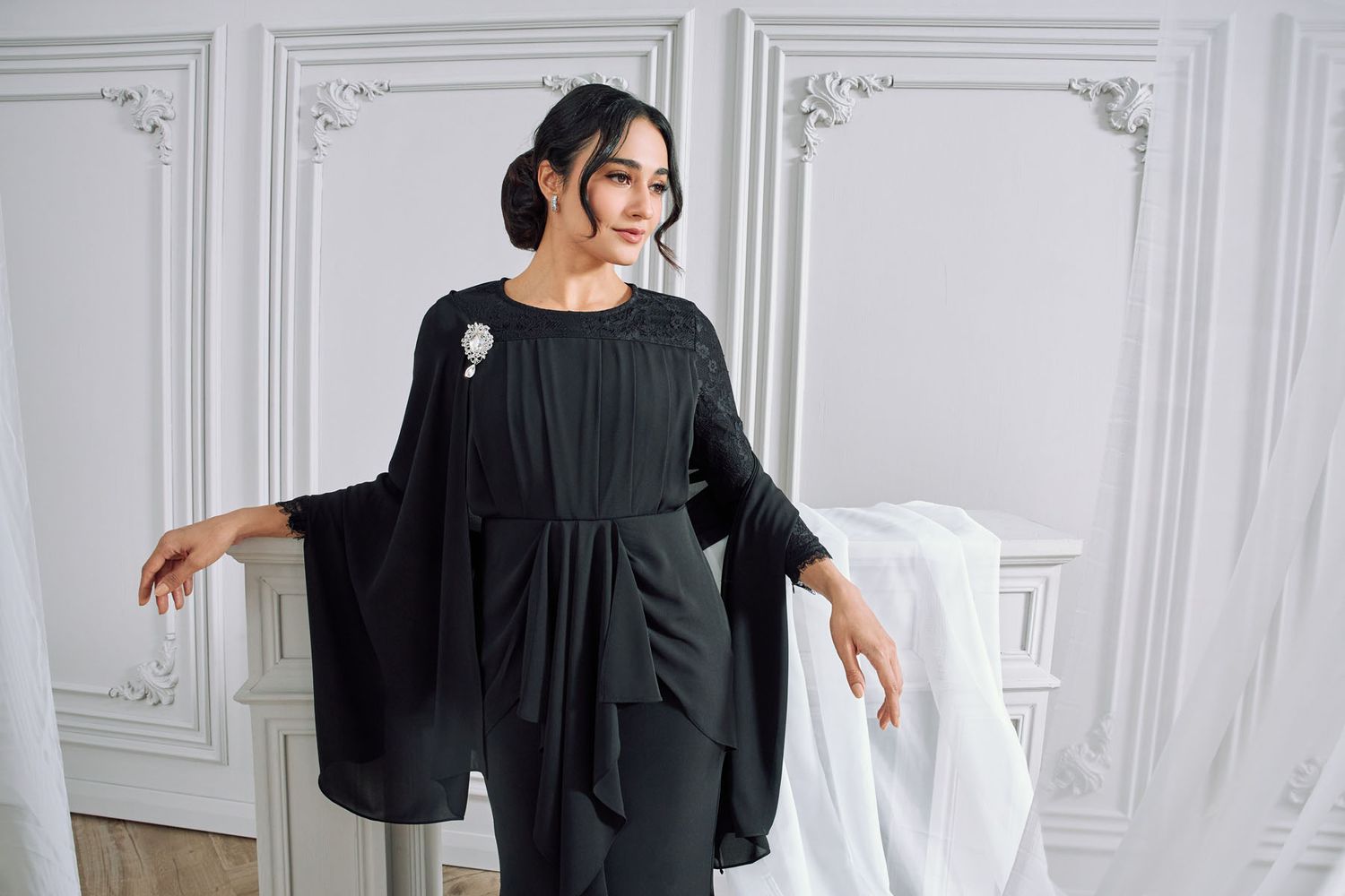 Woman in elegant black wide-sleeve modest jumpsuit with brooch posing in a bright white room.