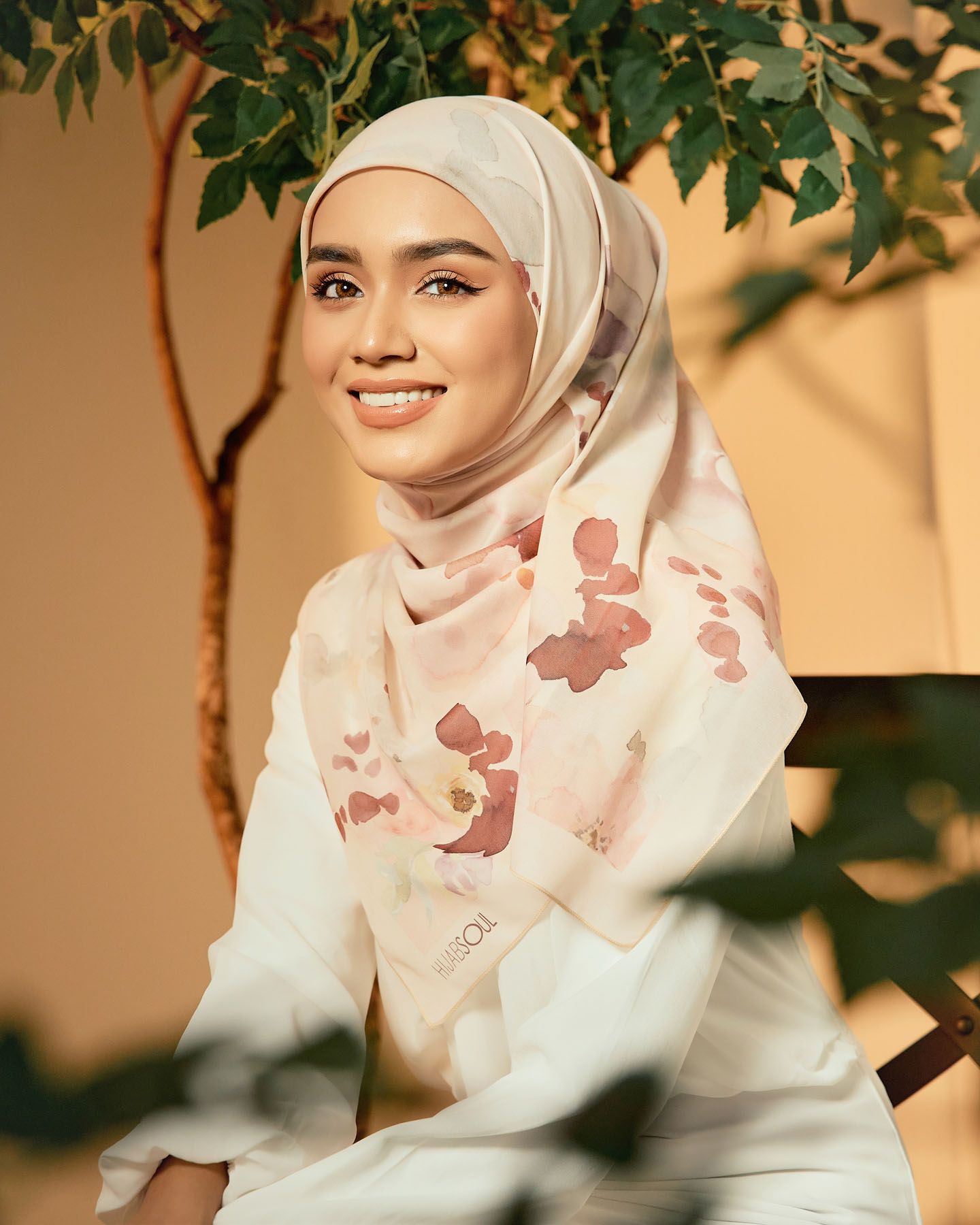 Smiling woman in a cream floral watercolor hijab seated near lush green foliage.
