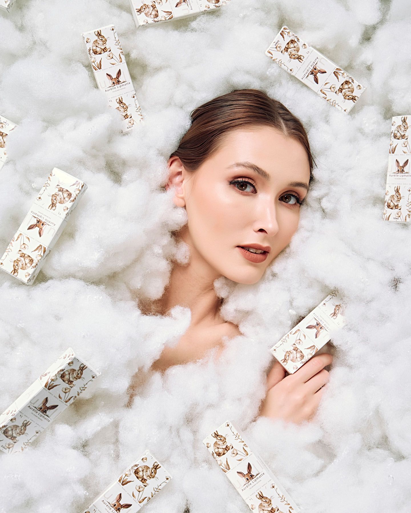 A model surrounded by white fluffy clouds and branded cosmetic packaging in a dreamy product shoot.