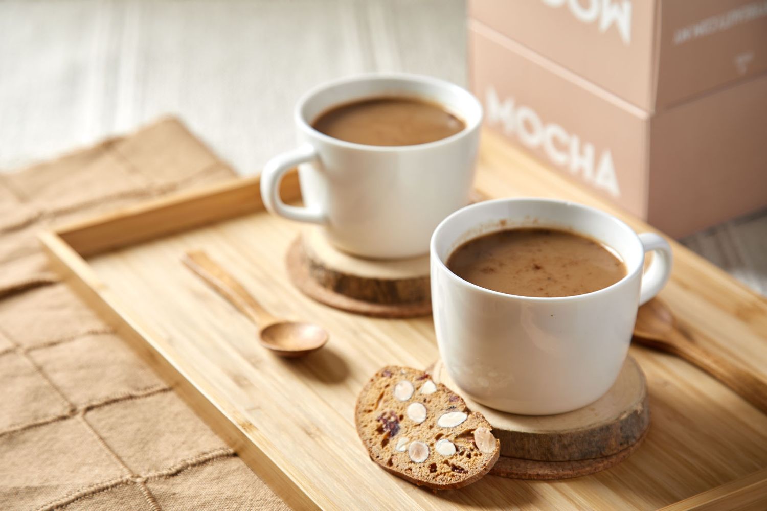 Two white mugs of mocha coffee on a wooden tray with an oat cookie and Mocha brand packaging.