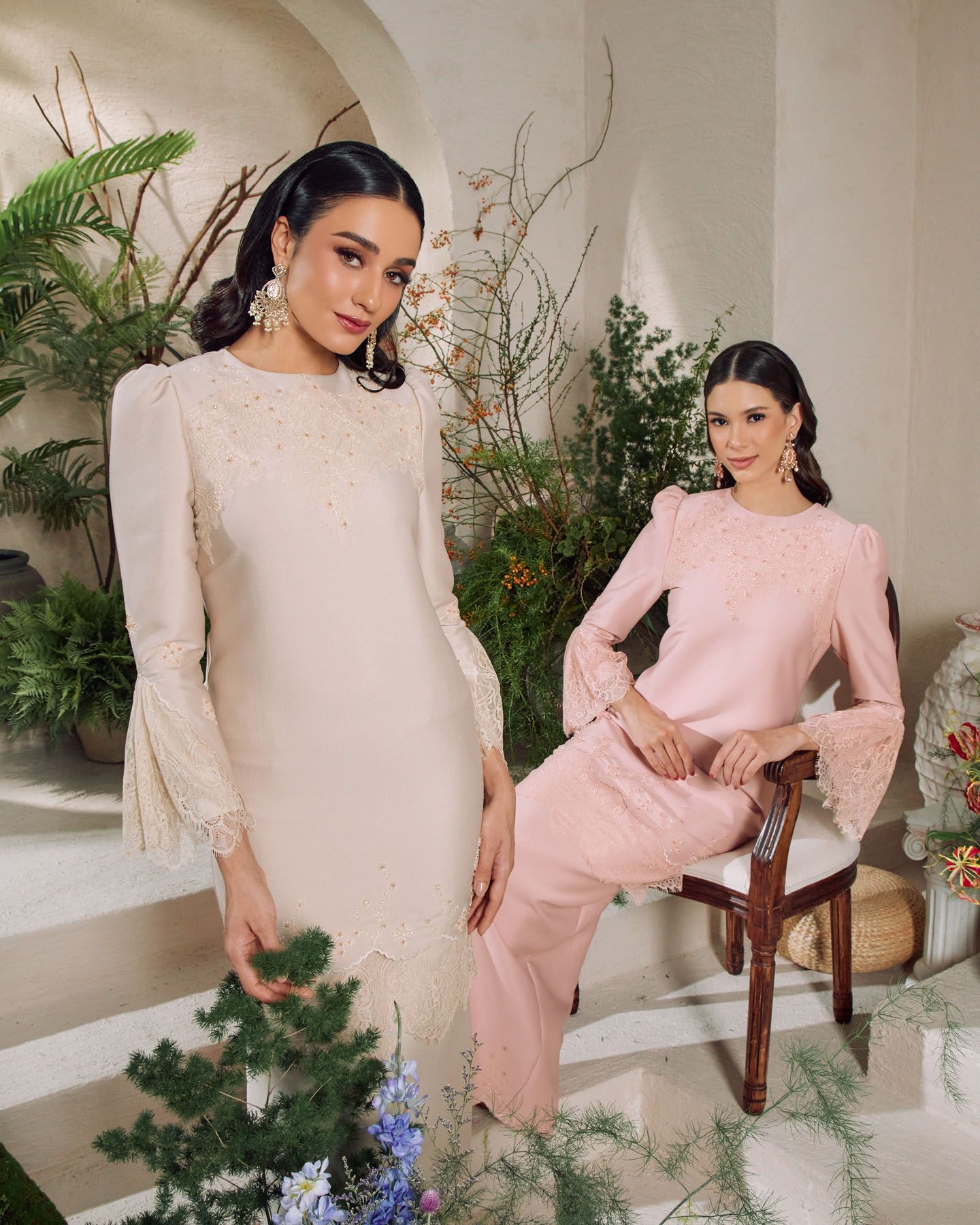 Two women model elegant baju kurung in cream with lace trim and blush pink with embroidery in a floral studio setting.