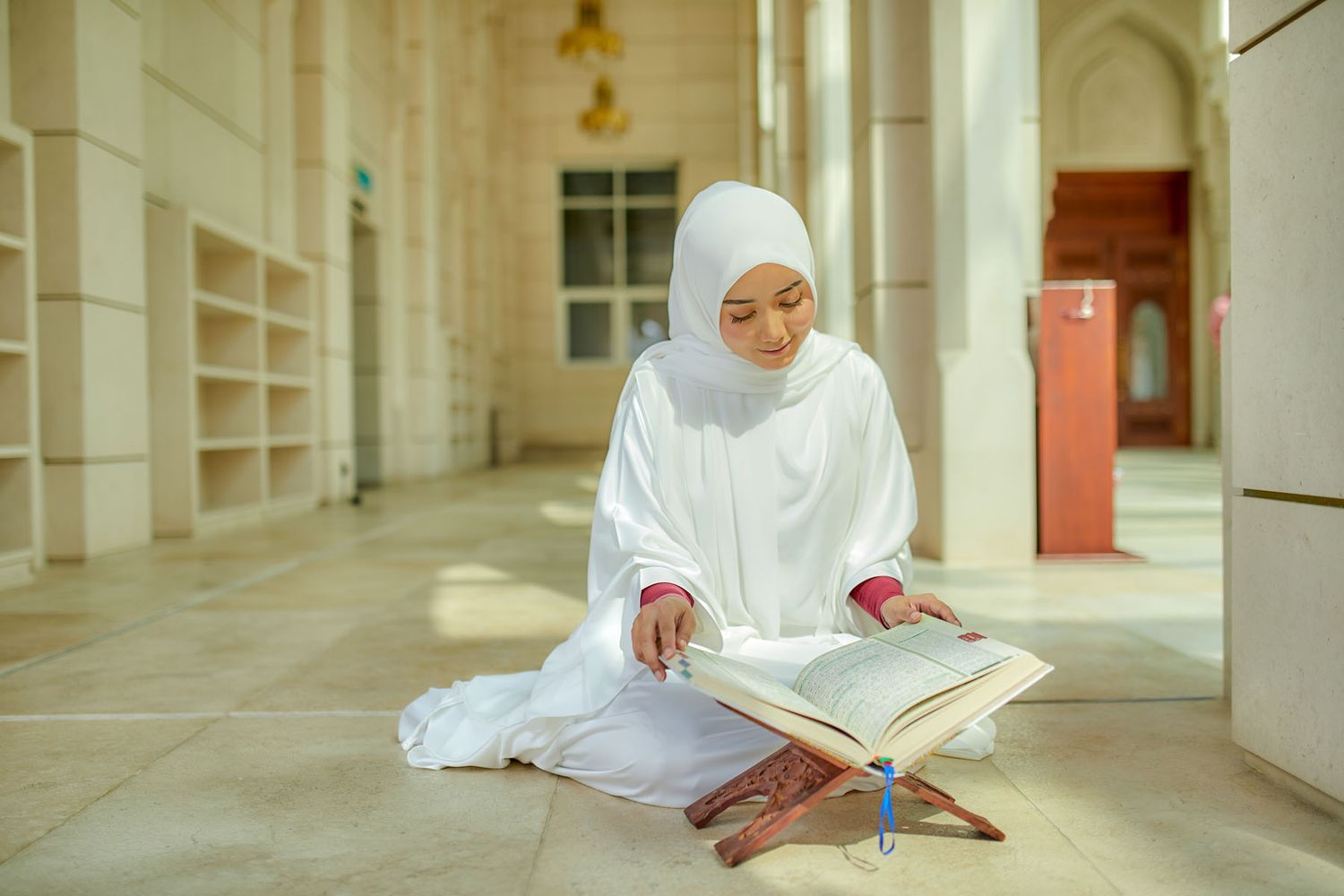 Young Muslim woman in white hijab sitting on floor reading Quran inside a mosque.