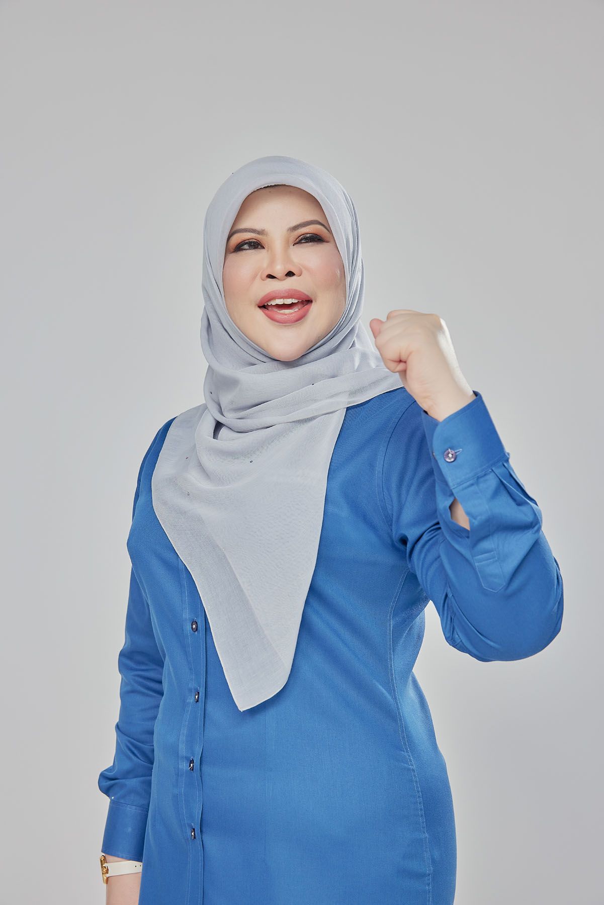 Confident Muslim woman in blue hijab outfit raising fist in victory gesture against grey background.