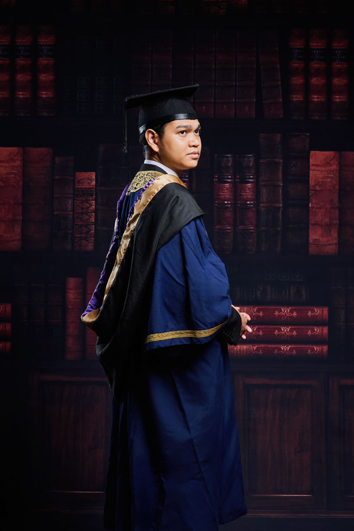 Male graduate in blue academic robe and cap posing sideways against dramatic dark bookshelf studio backdrop.
