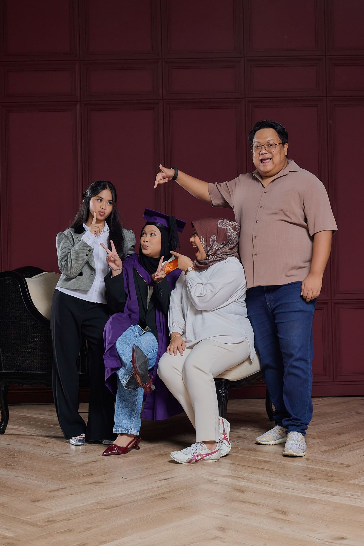 Family group posing playfully with graduate in purple cap and gown in red-walled studio.