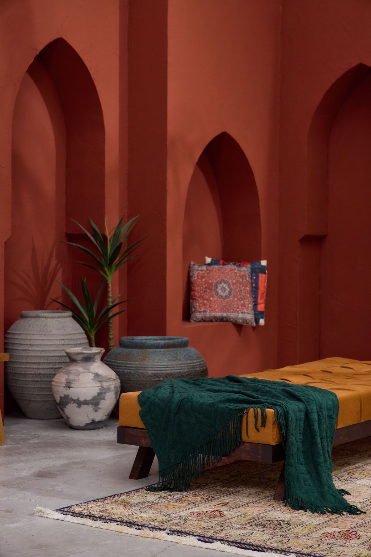 Moroccan interior with terracotta arched walls, large ceramic urns, mustard daybed with green throw, and Persian rug.