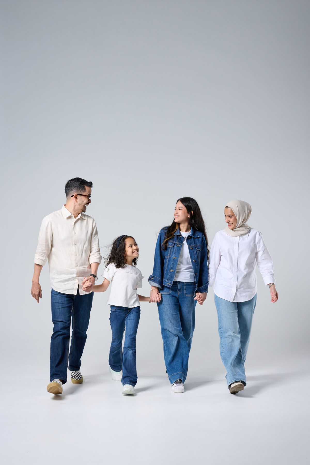 A happy Muslim family of four walking together holding hands, wearing casual denim outfits against a gray background.