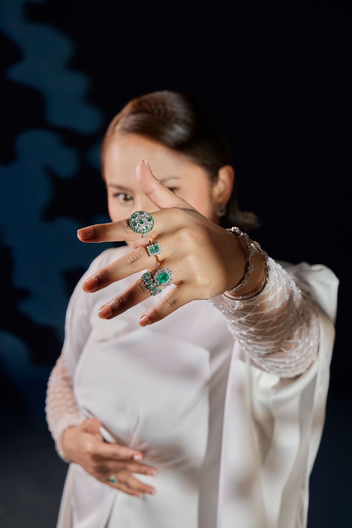 Woman in white outfit extending hand toward camera displaying emerald gemstone rings on dark background.