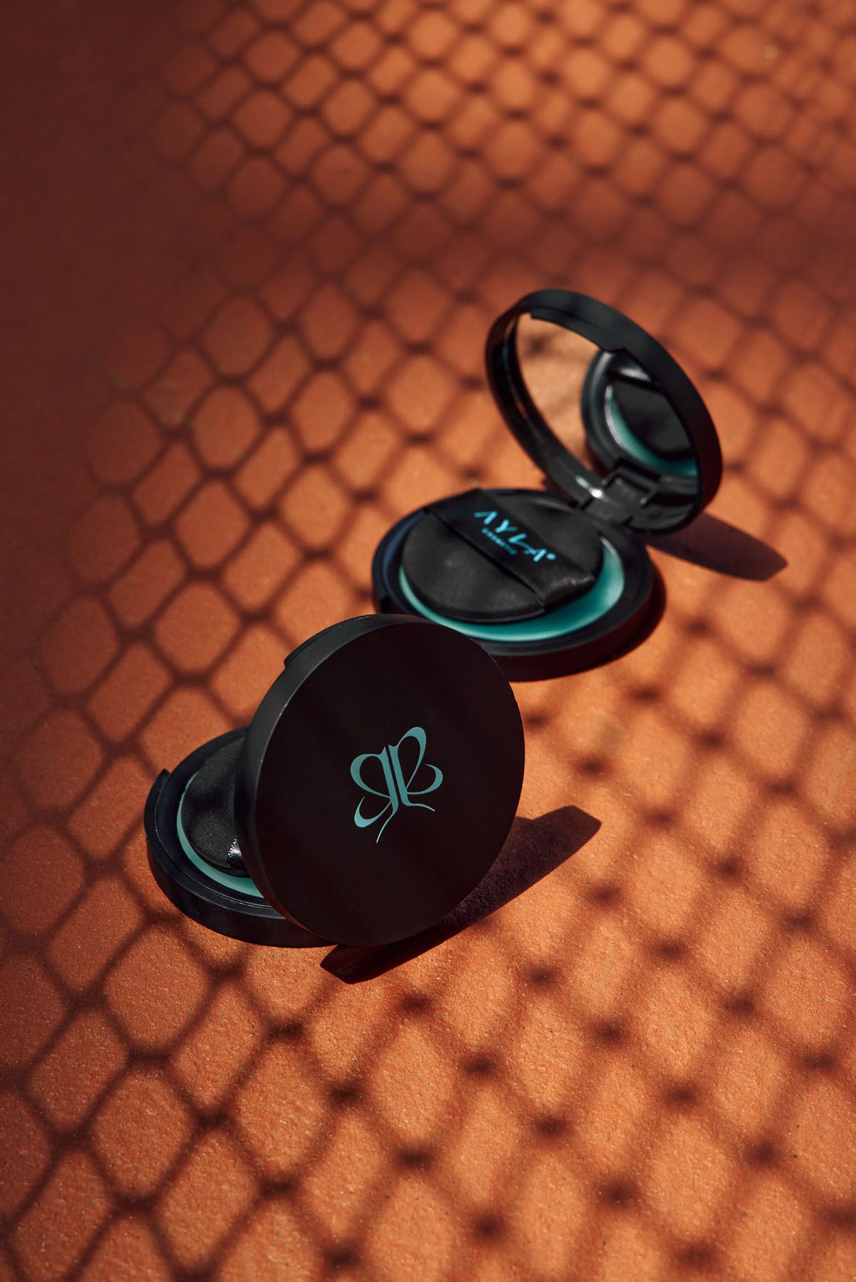Open black AYLA cosmetics compact powder with teal product and mirror, on terracotta surface with net shadow.