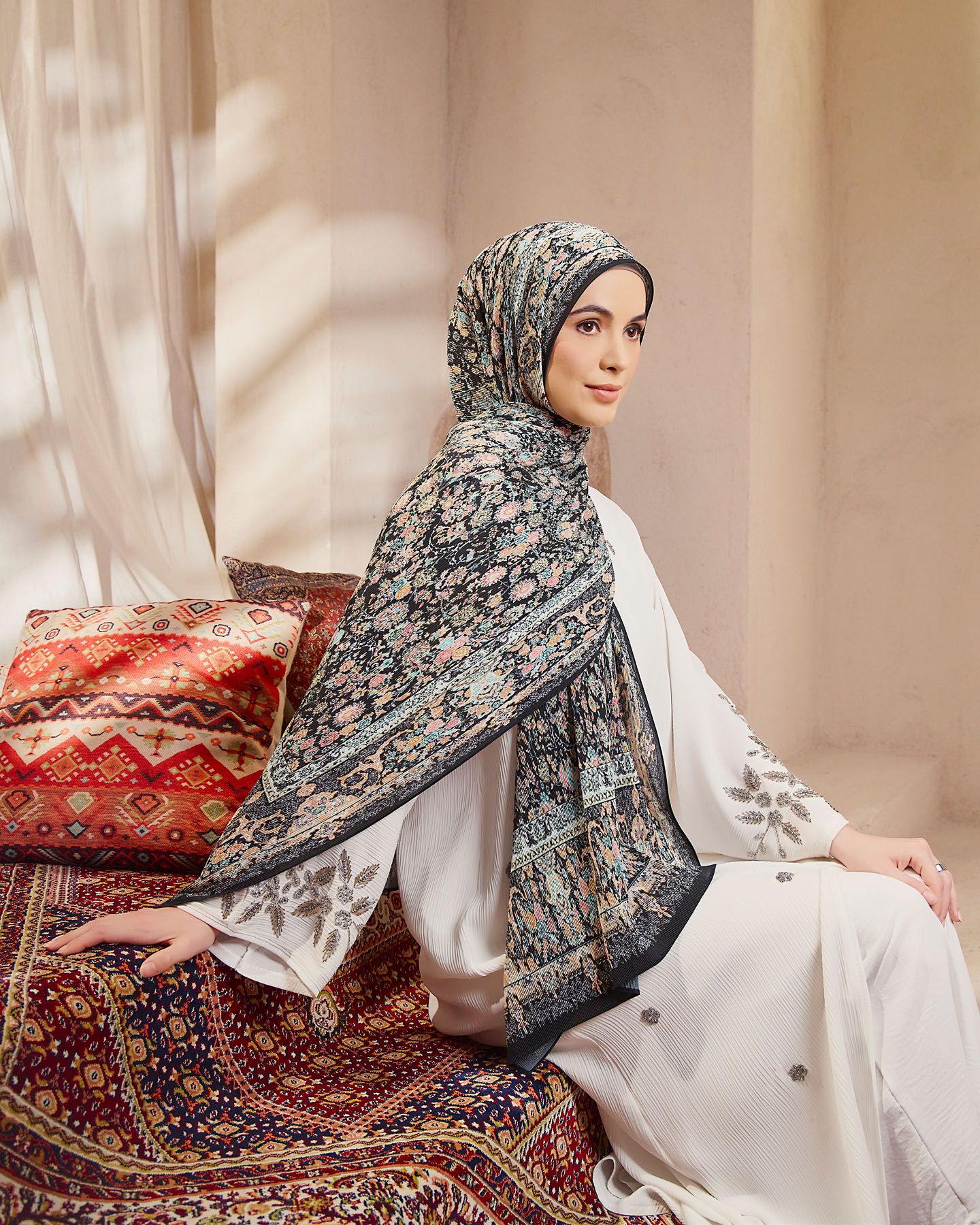 Woman wearing dark floral patterned hijab with white embroidered outfit seated on ornate carpet.
