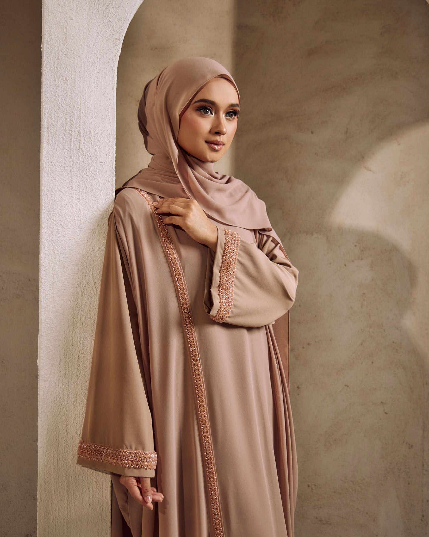 Woman in taupe hijab and embroidered abaya poses against stone wall.