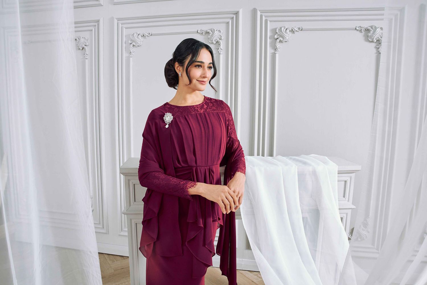Woman in deep burgundy modest dress with chiffon overlay and floral brooch in ornate white room.