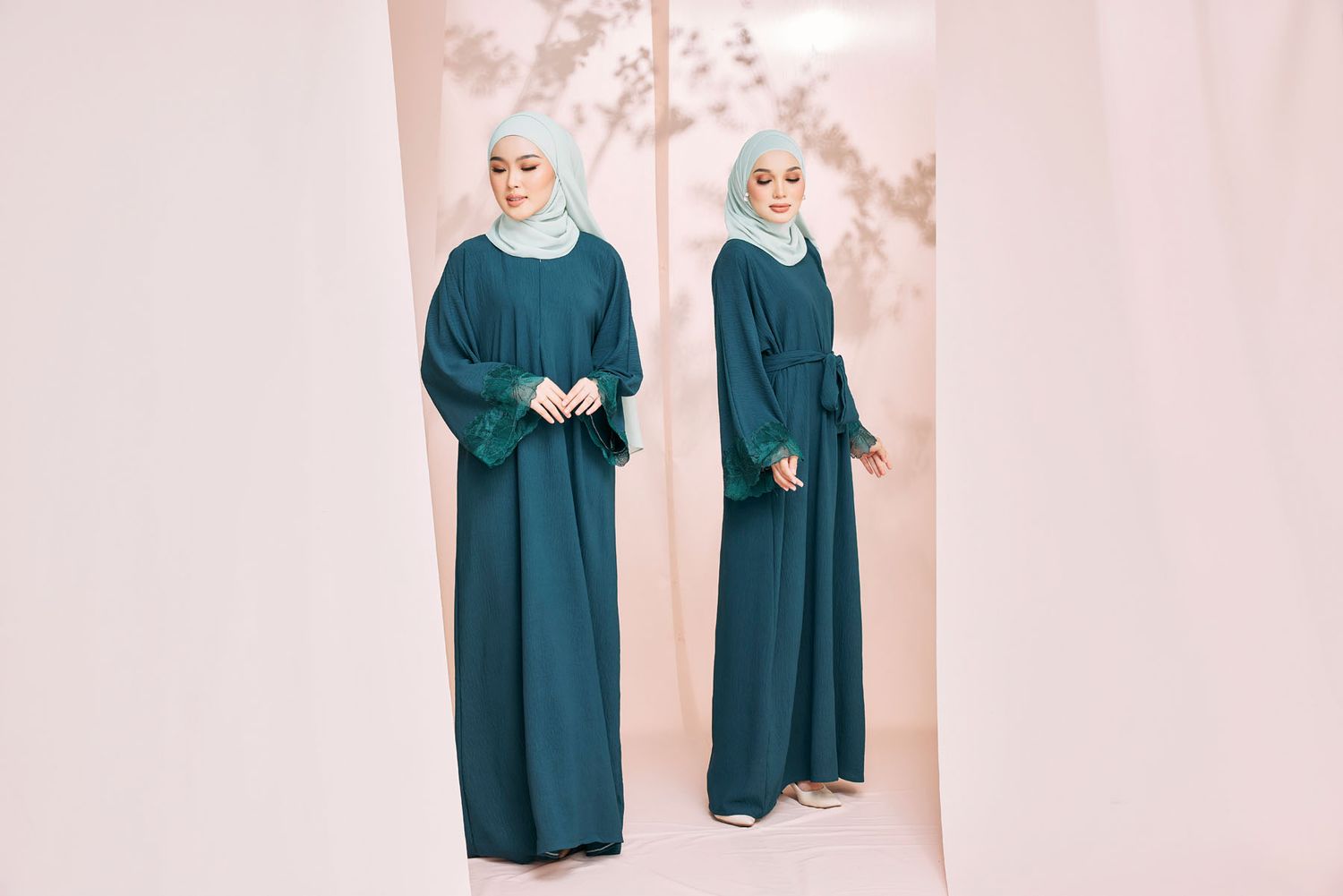 Two women in matching teal hijab and abaya with lace cuffs pose in a soft pink studio.