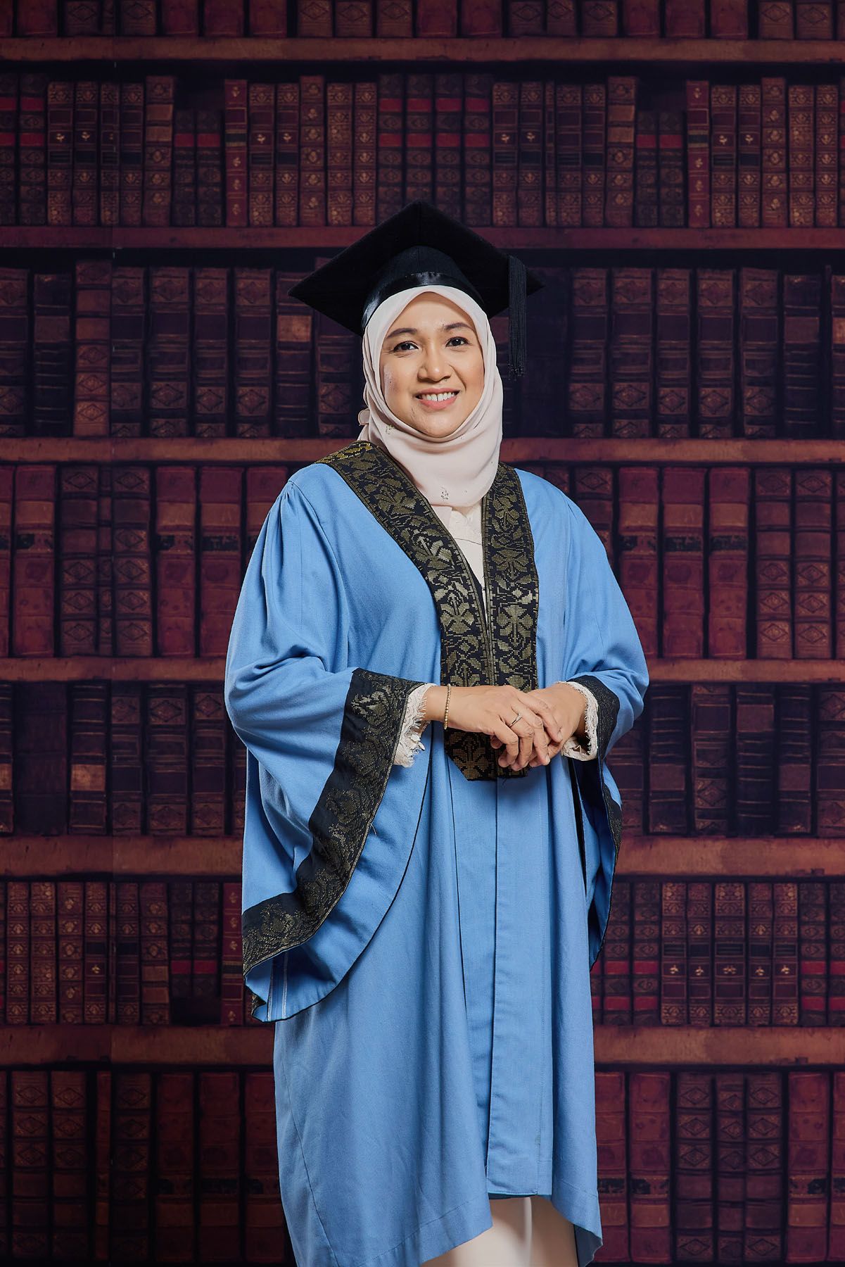 Smiling Muslim woman in light blue graduation gown and hijab standing before decorative bookshelf background.