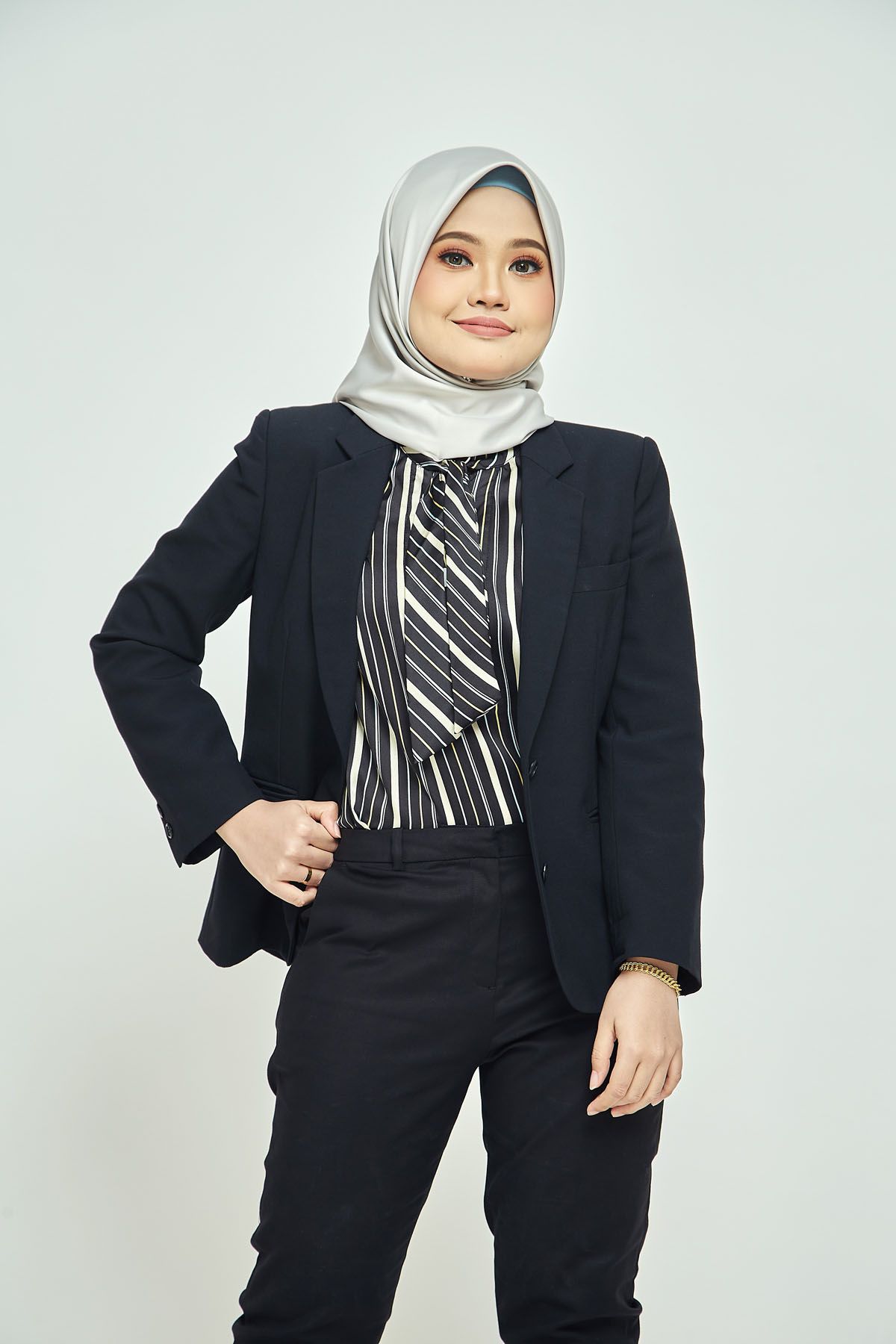 Young Muslim woman in black blazer with striped top and grey hijab posing on white background.