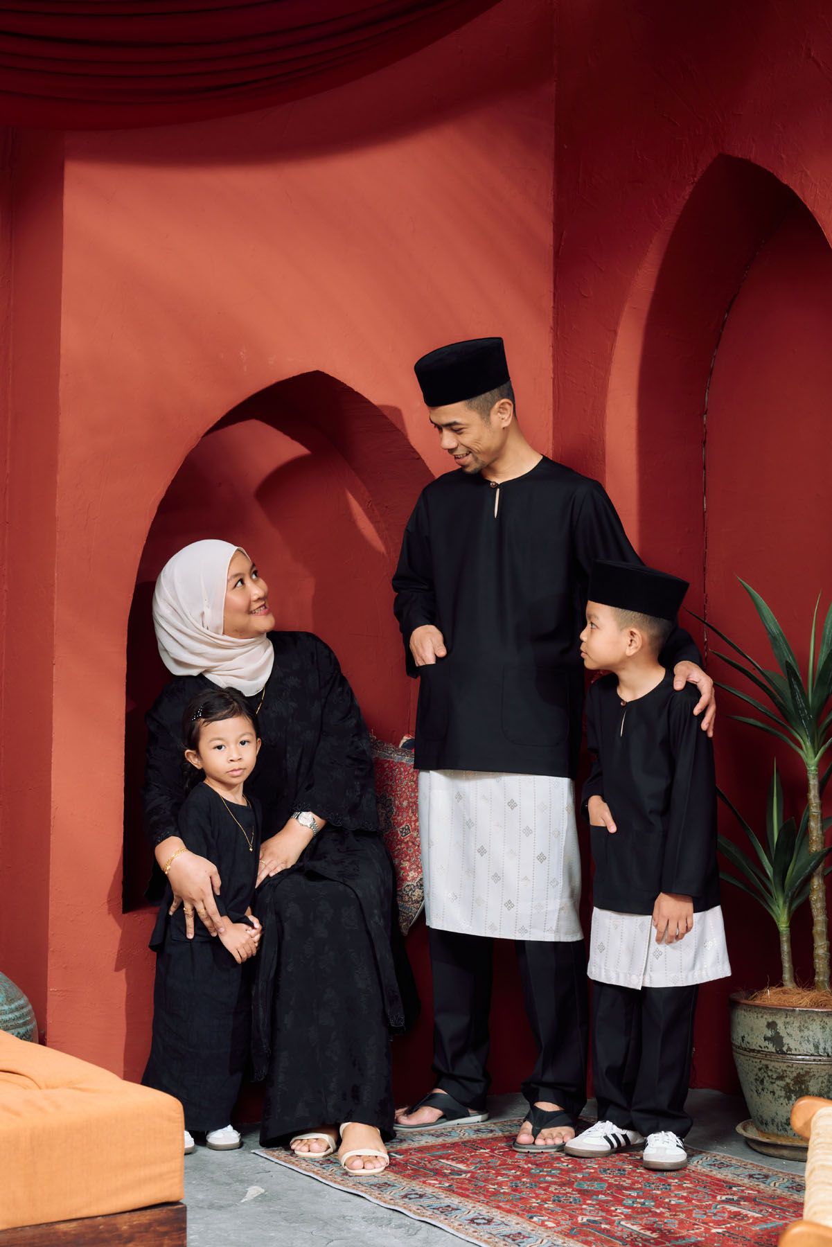 Muslim family in matching black traditional Baju Raya outfits posing in red arched courtyard.
