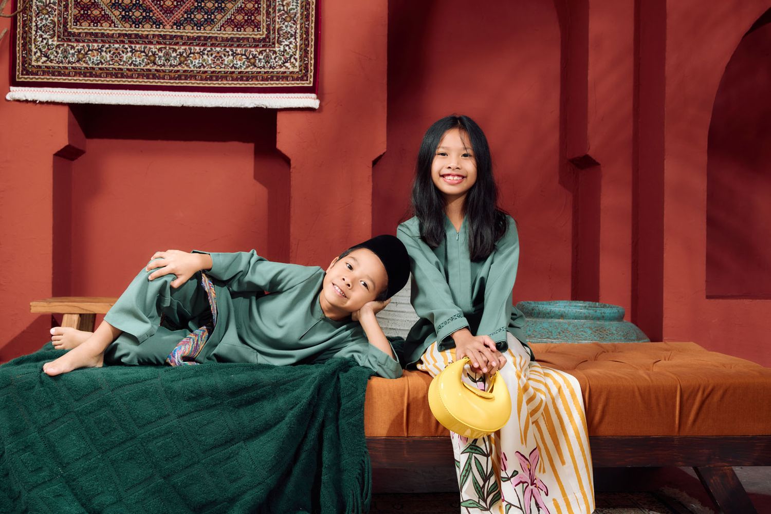 Two children in green Malay traditional wear posing on a bed with ornate carpets in a red studio setting.