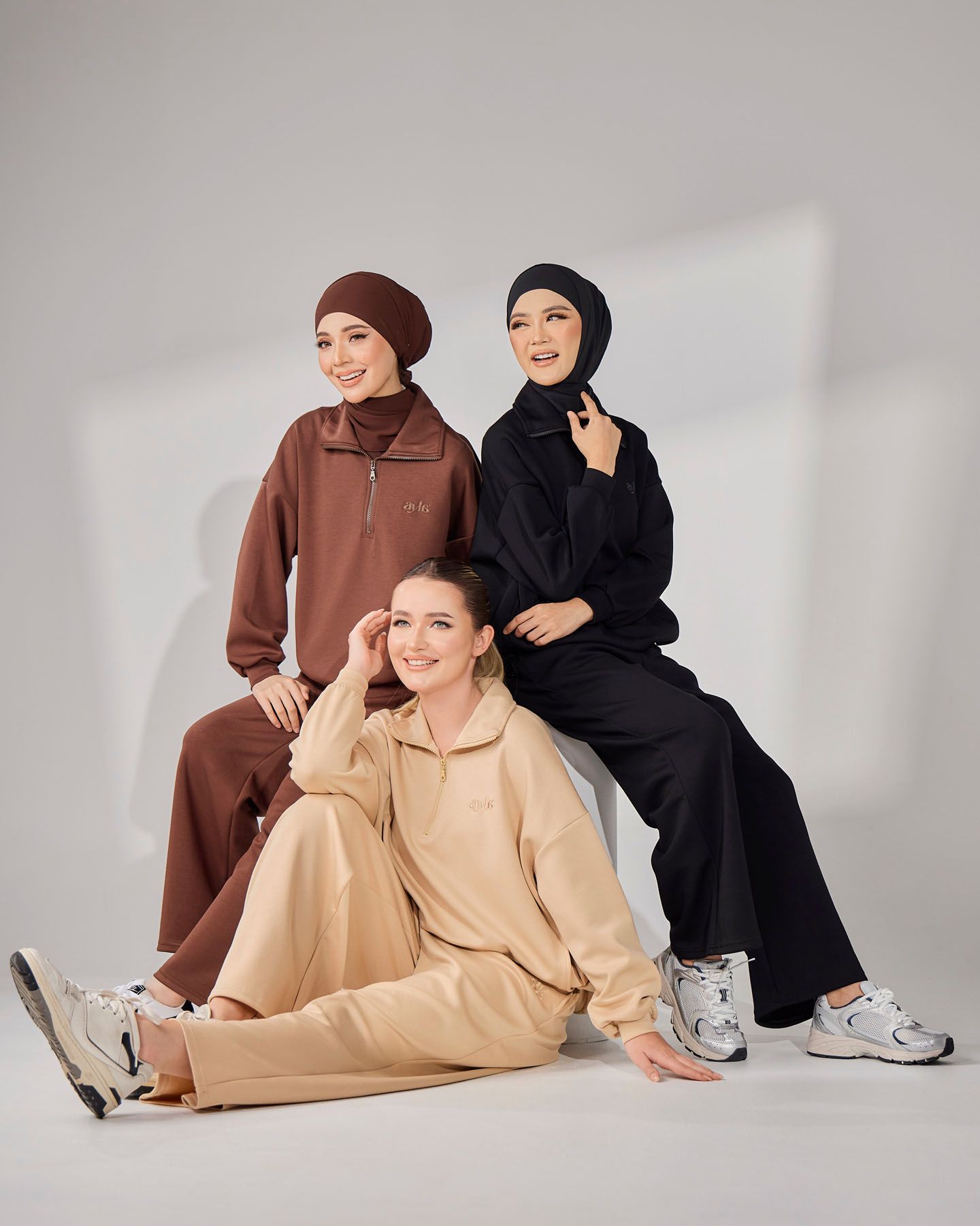 Three women in modest coordinating loungewear sets in black, brown, and beige with hijabs posing together.