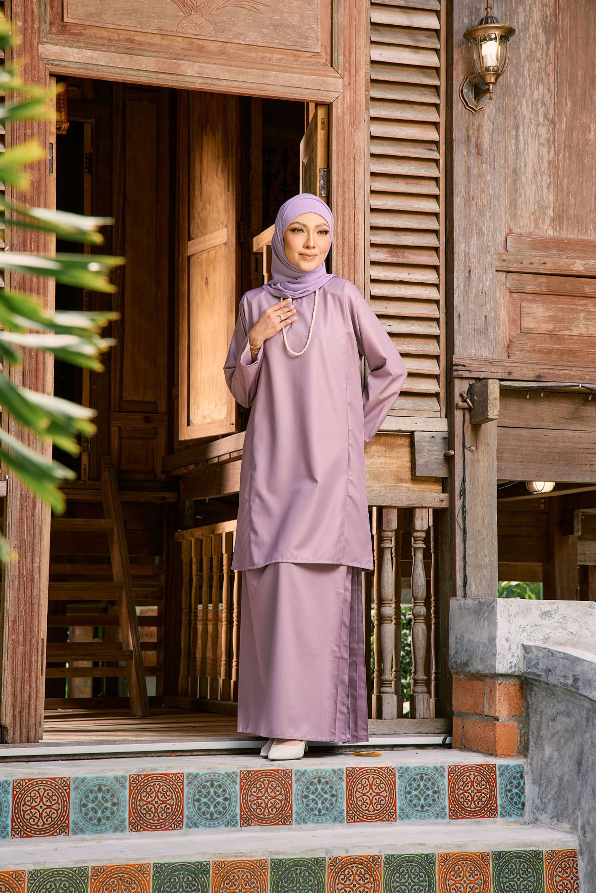 Woman in mauve purple baju kurung with matching hijab standing at entrance of traditional wooden house.