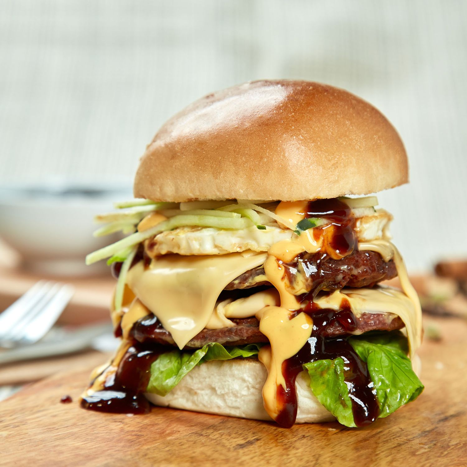 Gourmet double cheeseburger with lettuce, melted cheese, and drizzled BBQ sauce on wooden board.