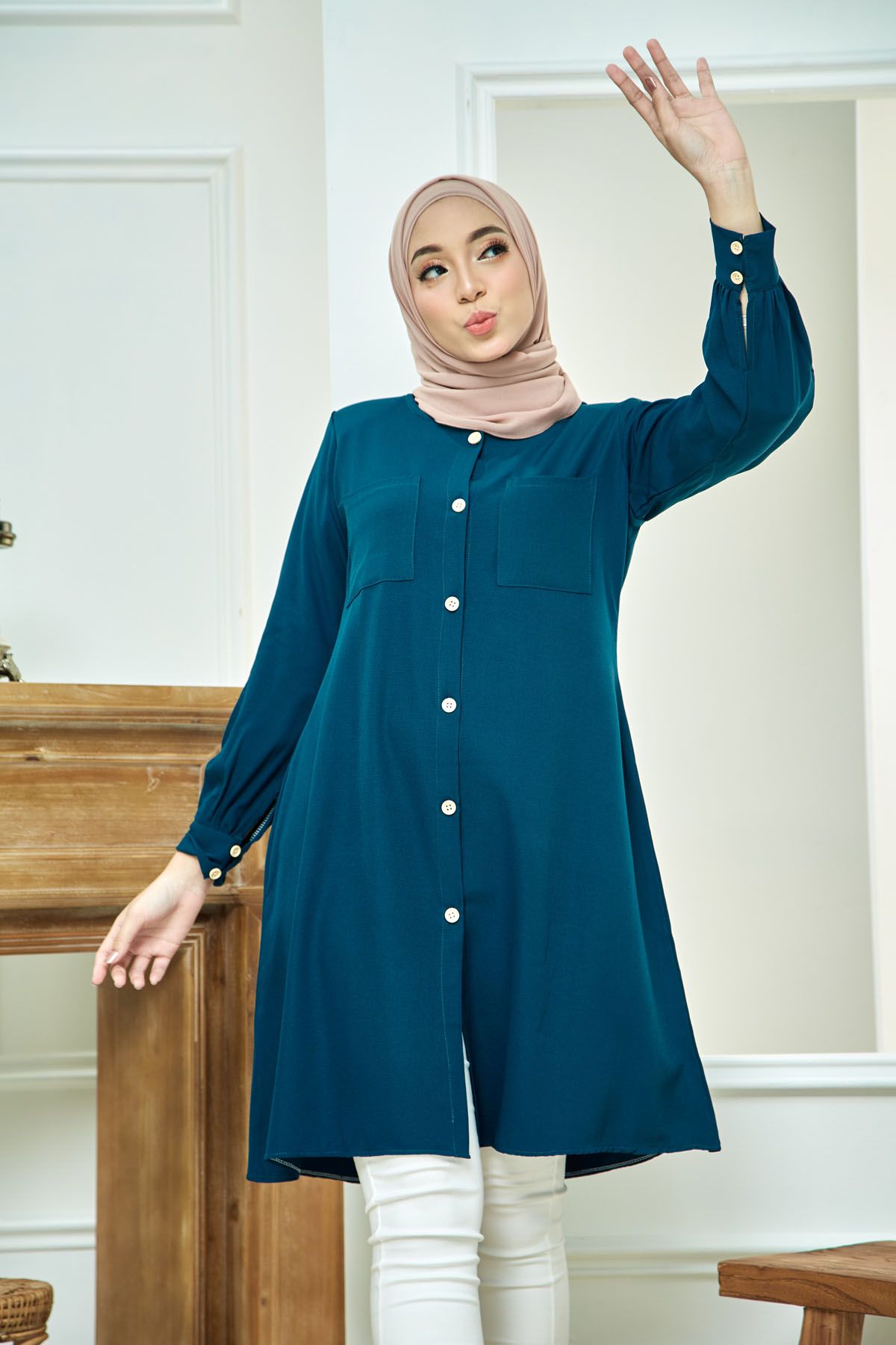 Woman in teal button-down tunic with white pants and beige hijab posing near wooden furniture.