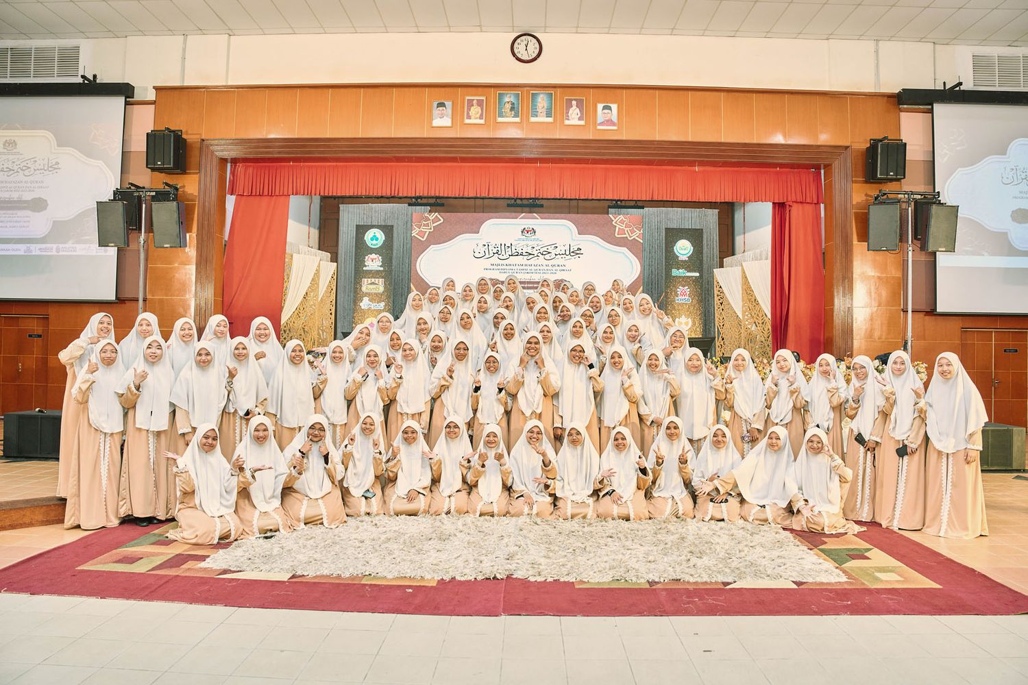 Large group of Muslim women in white hijabs pose together on stage at a formal Islamic graduation ceremony.
