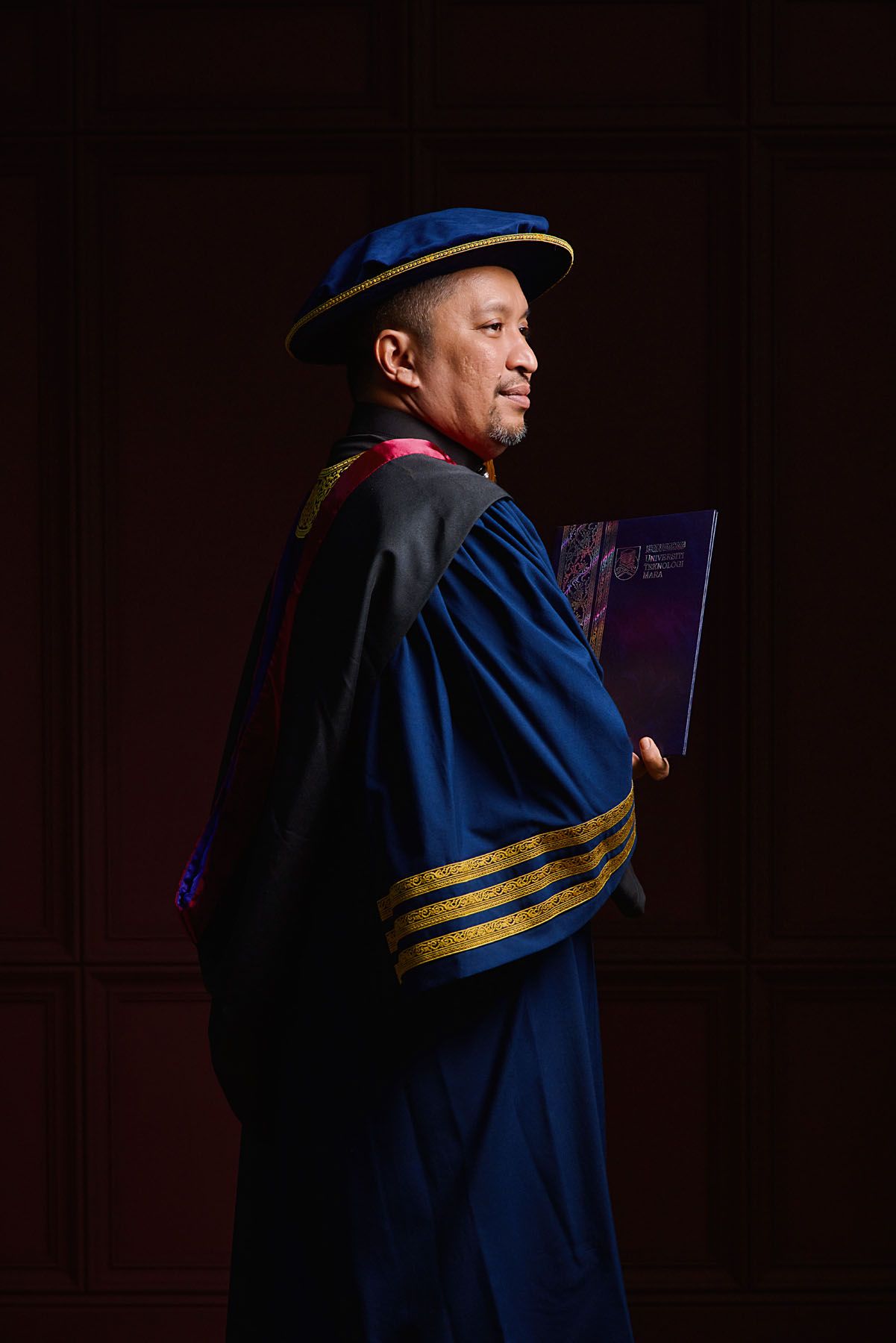 Academic professor in blue doctoral robes and cap holds book in dramatic side profile against dark background.