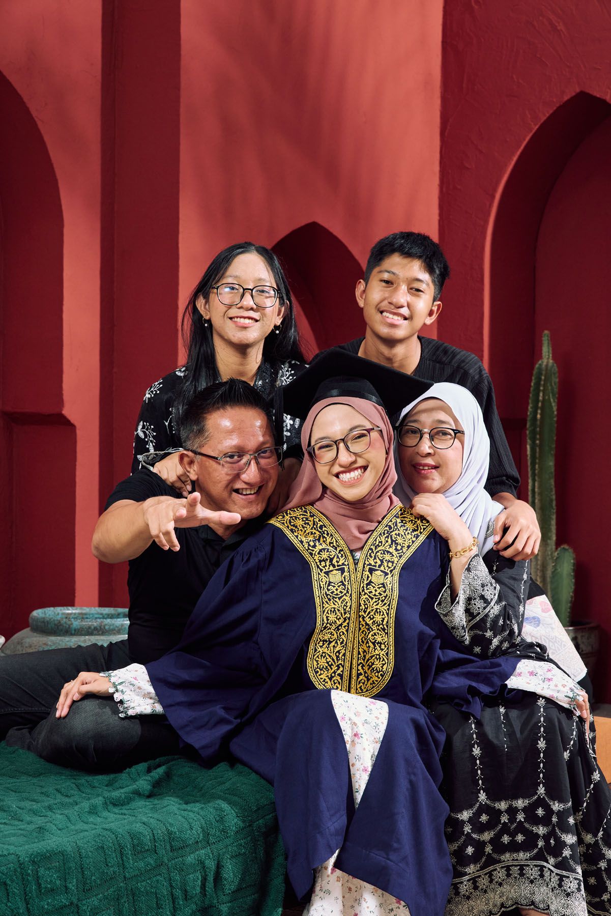 Happy Muslim graduate in navy gown celebrates with smiling family against a vibrant red arched backdrop.