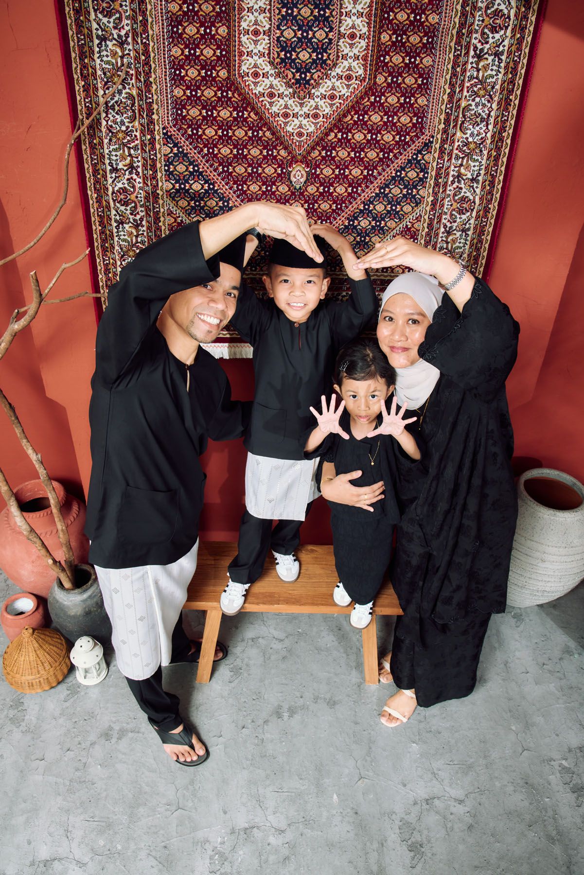 Muslim family in matching black outfits forming heart shape with hands before ornate carpet.