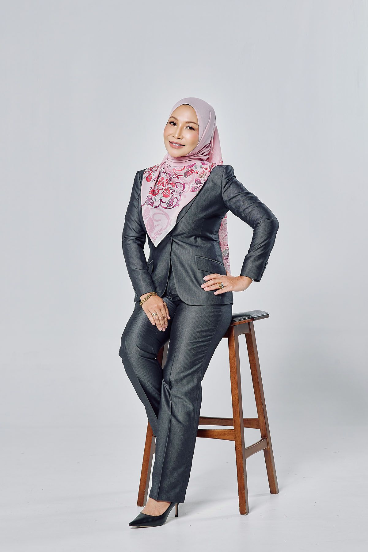 Professional Muslim woman in a pink hijab and gray business suit posing beside a wooden stool.