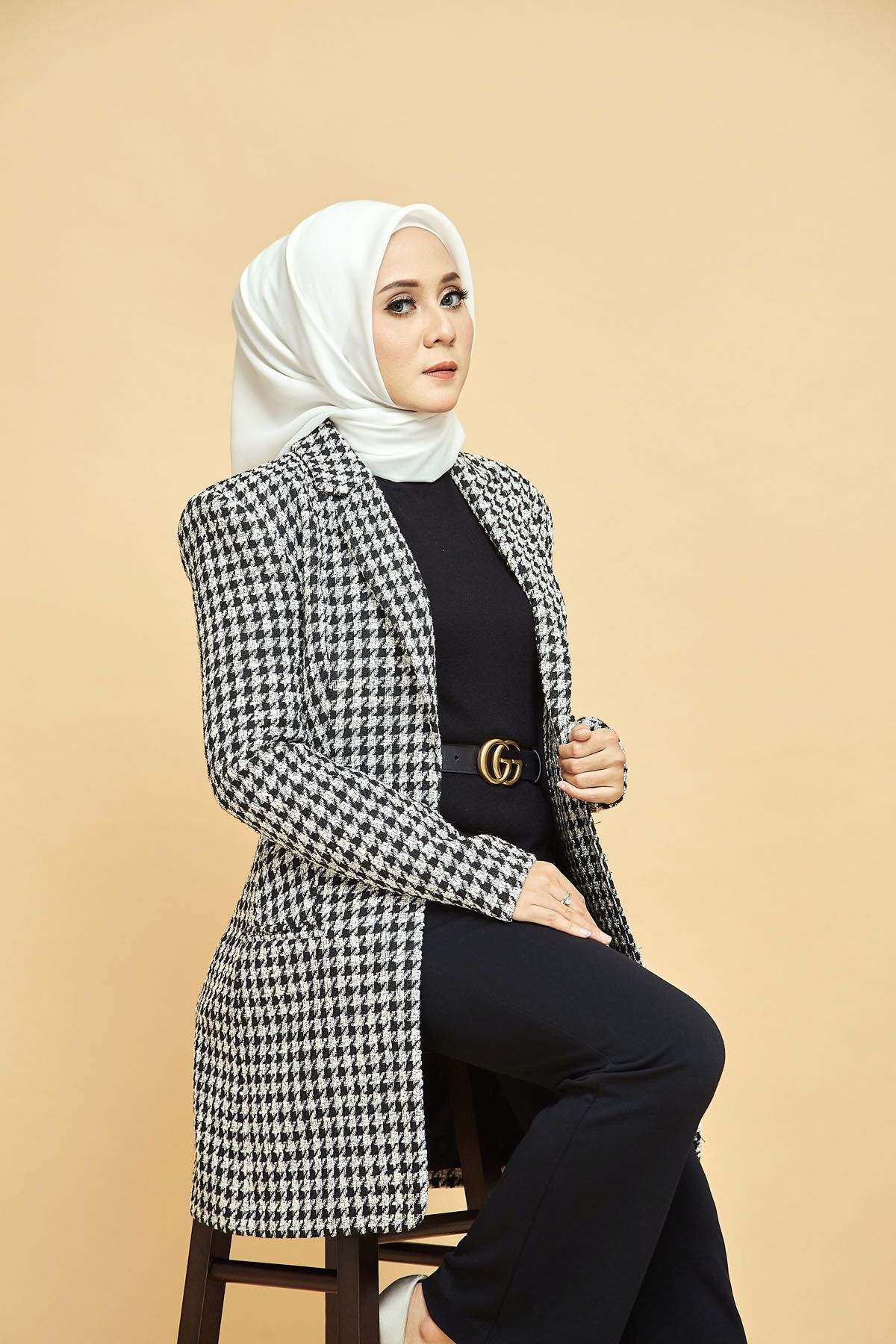 Muslim woman in white hijab wearing houndstooth blazer seated on stool against beige background.