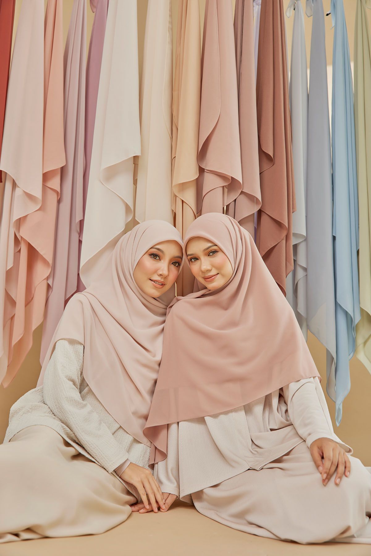 Two women in pastel pink hijabs sitting together before a colorful array of hanging hijab fabrics.