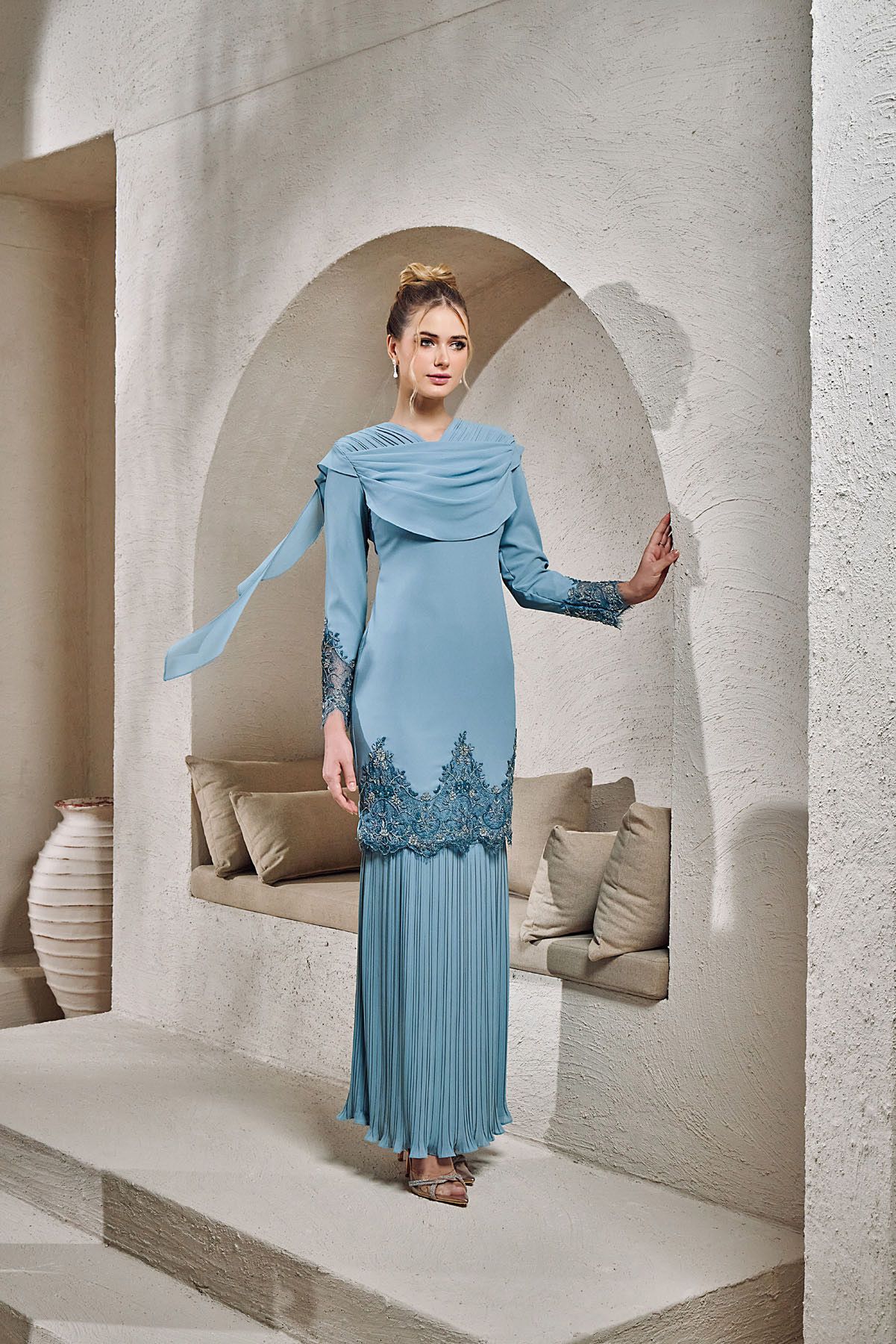 Elegant blue pleated baju kurung gown with lace hem detail and draped scarf in Mediterranean setting.