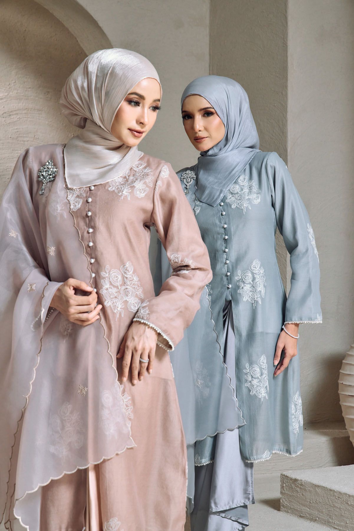 Two women in pastel pink and blue floral embroidered baju kurung with pearl buttons pose elegantly.