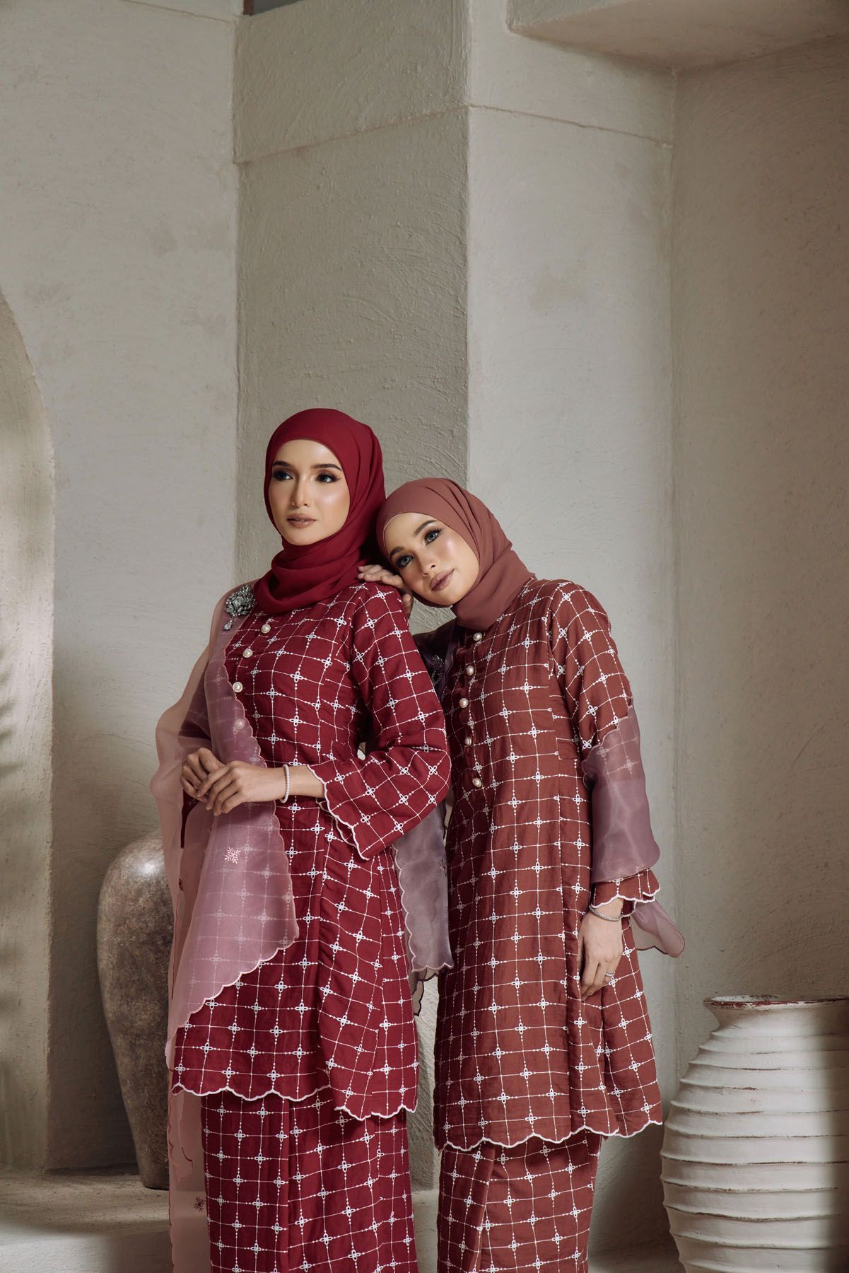 Two women in matching dark red checkered baju kurung sets with hijabs pose in architectural setting.